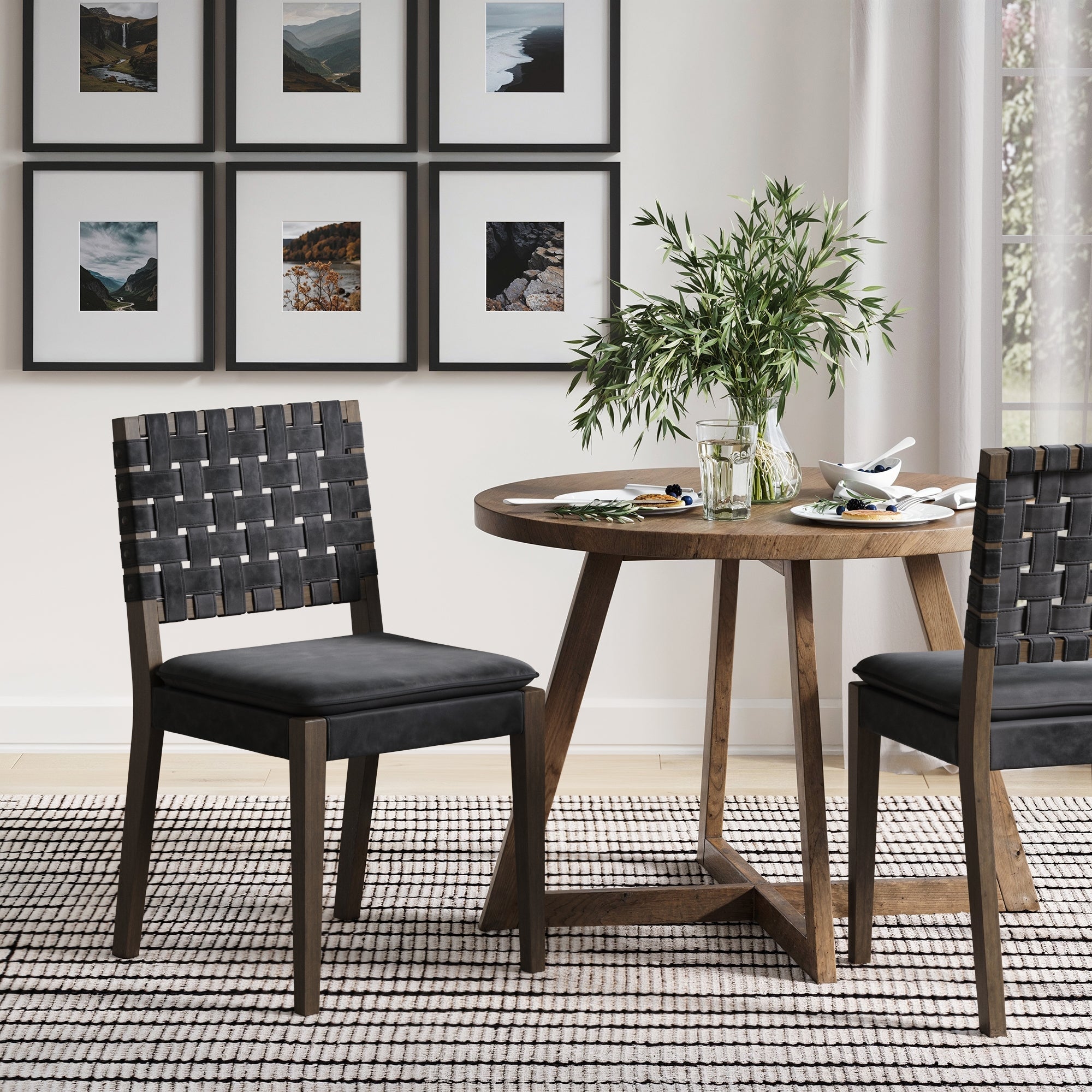 A round wooden table with two Nathan James Woven Faux Leather Dining Chairs in black sits on a patterned rug. The table holds a glass vase with greenery and dishes, while framed landscape photos hang on the white wall behind.