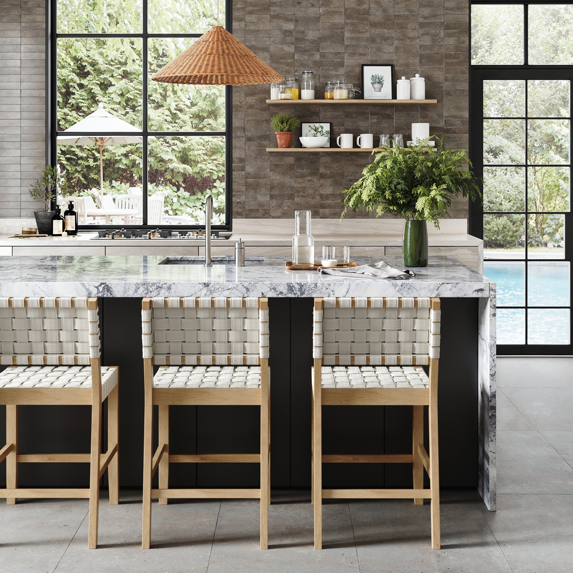A modern kitchen features a marble island, three Nathan James Faux Leather Woven Counter Height Bar Stools in off white, green foliage in a vase, open shelves with dishes, large windows, and views of the pool and garden.
