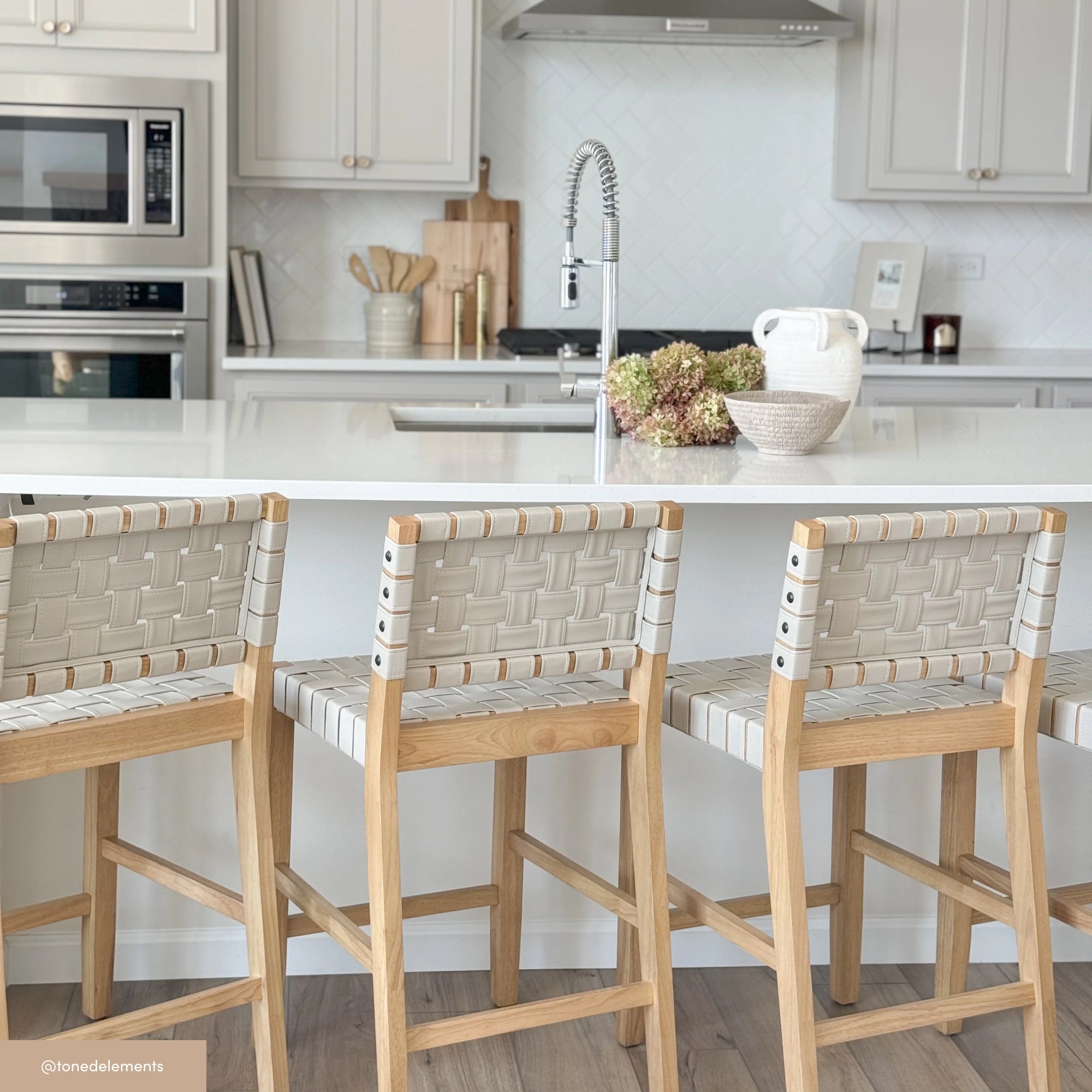 A modern kitchen with a white island, three woven-back wooden barstools, a stainless steel oven and microwave, and neutral-toned decor, including a bowl of flowers and a white jug on the countertop.