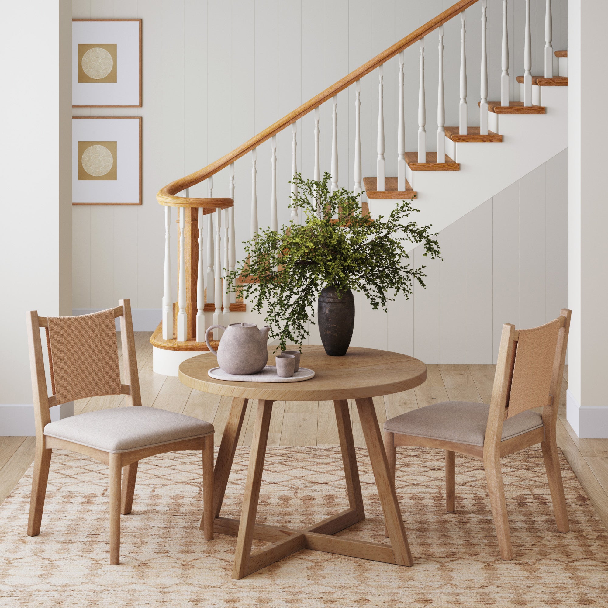 A round wooden table with two Nathan James Arched Back Seagrass Dining Chairs (set of 2), plus a vase with green foliage and ceramics, sits on a patterned rug before a staircase in a bright, modern room.