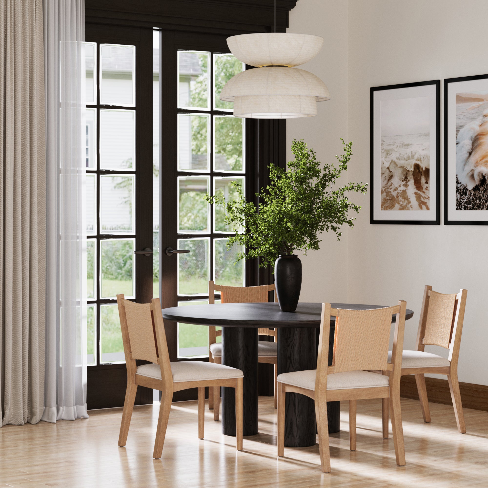 A modern dining room with a round black table, four Nathan James Arched Back Seagrass Dining Chairs in light brown, a black vase with green branches, large curtained windows, and two framed nature photos on the wall.