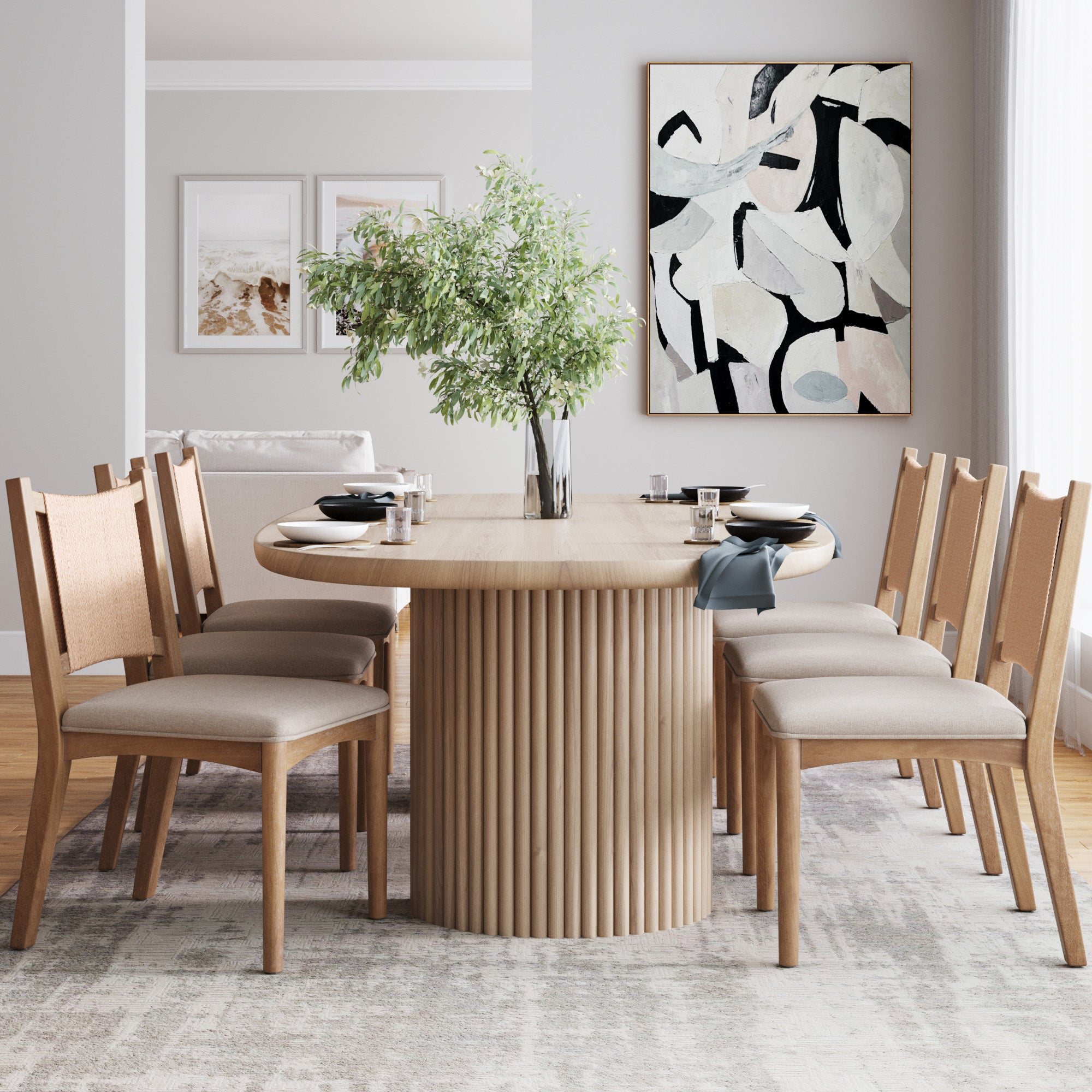 A modern dining room with an oval table, six Nathan James Arched Back Seagrass Dining Chairs in light brown, a green leafy centerpiece, neutral wall art, soft natural lighting, light walls, and a gray patterned rug.