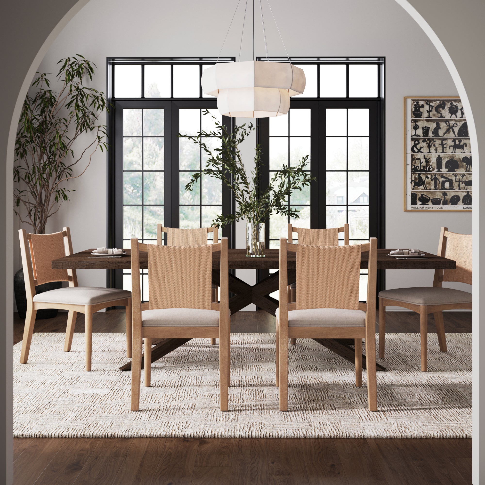 A modern dining room with a solid wood table, six Nathan James Arched Back Seagrass Dining Chairs in light brown, a light rug, bright windows, round white chandelier, green plants, black-framed art, and a tall plant for an airy feel.