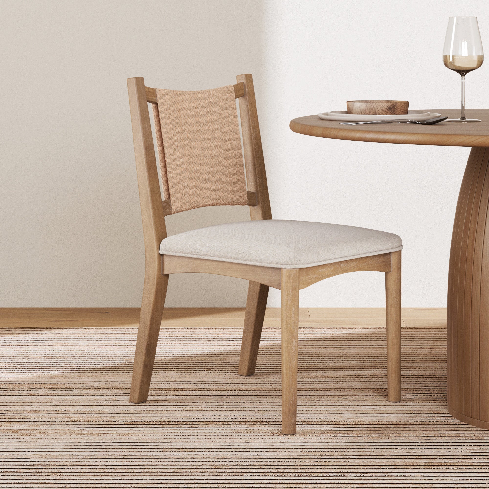 The Nathan James Arched Back Seagrass Dining Chair in light brown features a solid wood frame, light fabric seat, and synthetic seagrass backrest, placed by a round wooden table set with dishes and a wine glass on a striped rug in a minimalist room.