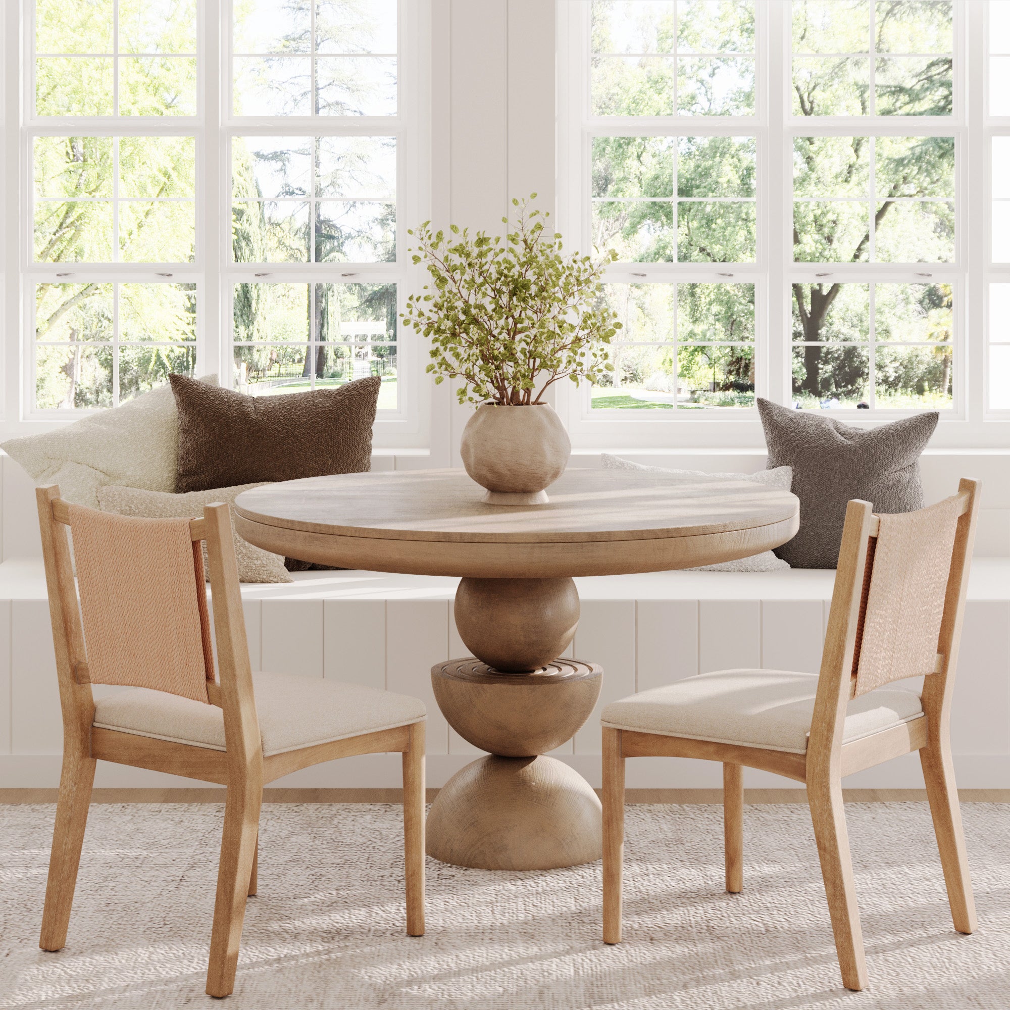 A round wooden dining table with a vase of greenery sits between two Nathan James Arched Back Seagrass Dining Chairs in light brown, featuring solid wood frames and synthetic seagrass backrests. A window seat behind displays neutral throw pillows.
