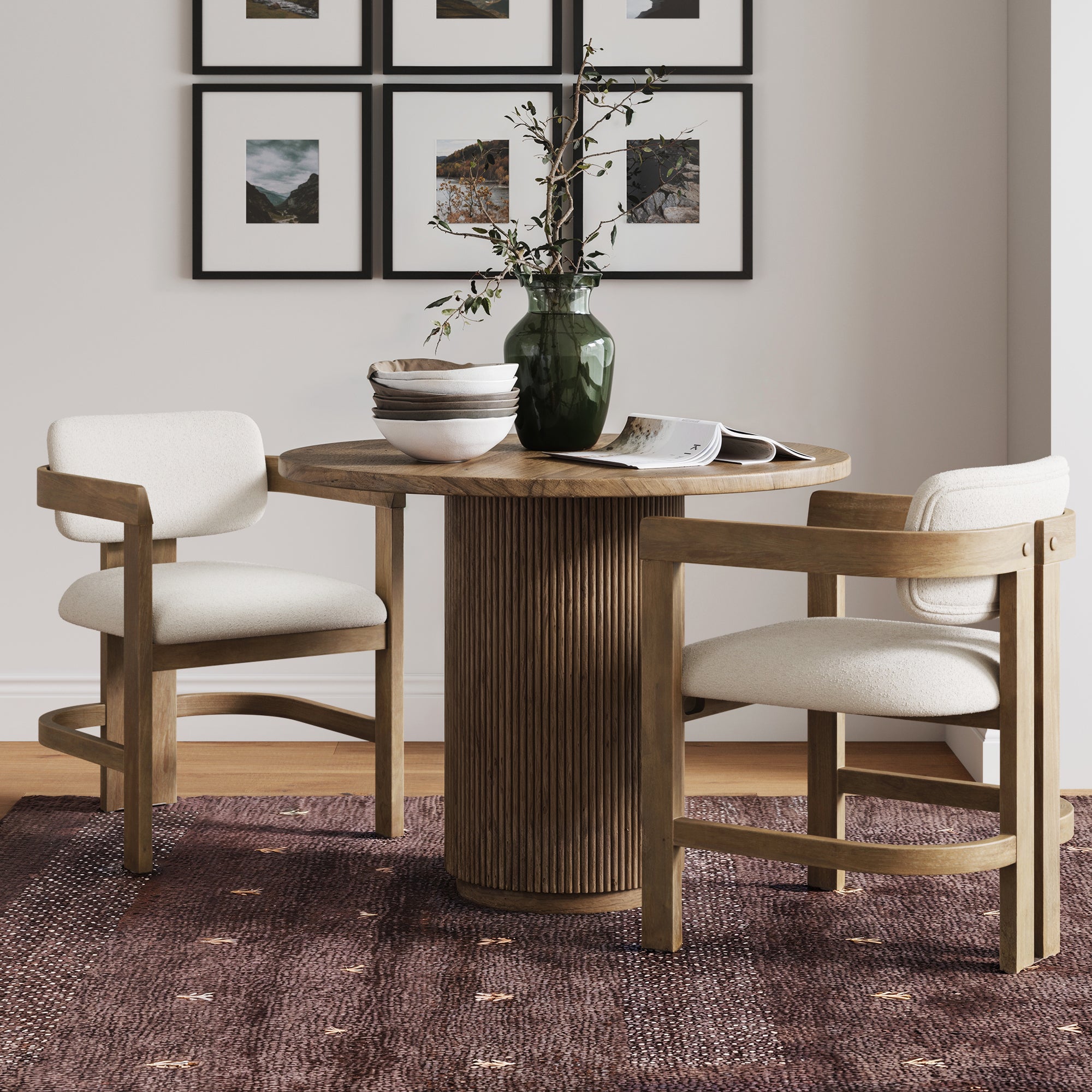 A round wooden table with two Nathan James Boucle Dining Chairs with curved backs and arms in light brown, a vase with branches, and bowls sits on a purple patterned rug. Framed landscape photos hang on the wall behind.