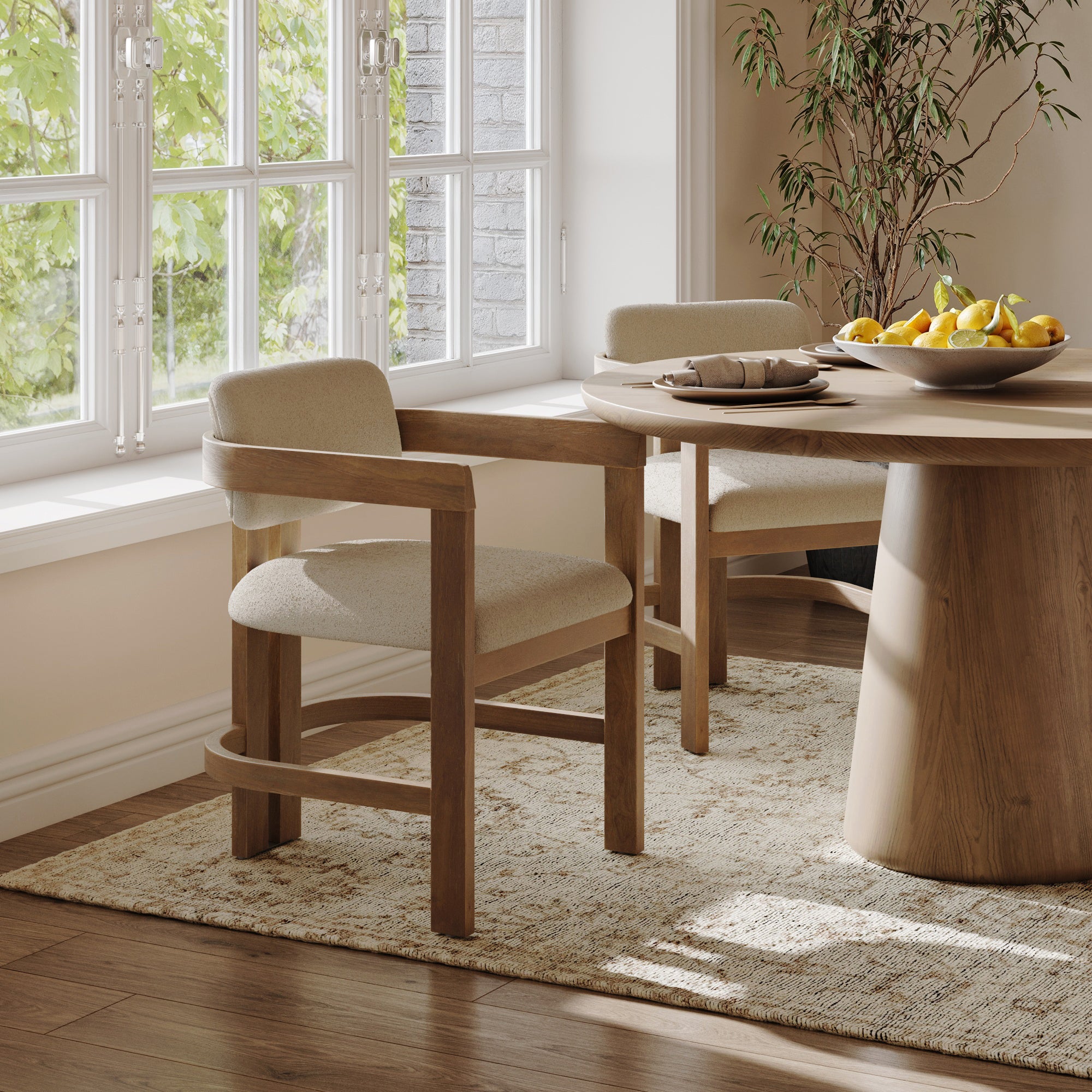 Two Nathan James Boucle Dining Chairs with curved backs and arms in light brown sit at a round wooden table set with fruit and plates. Sunlight streams through large windows, illuminating the cozy room with a beige rug.