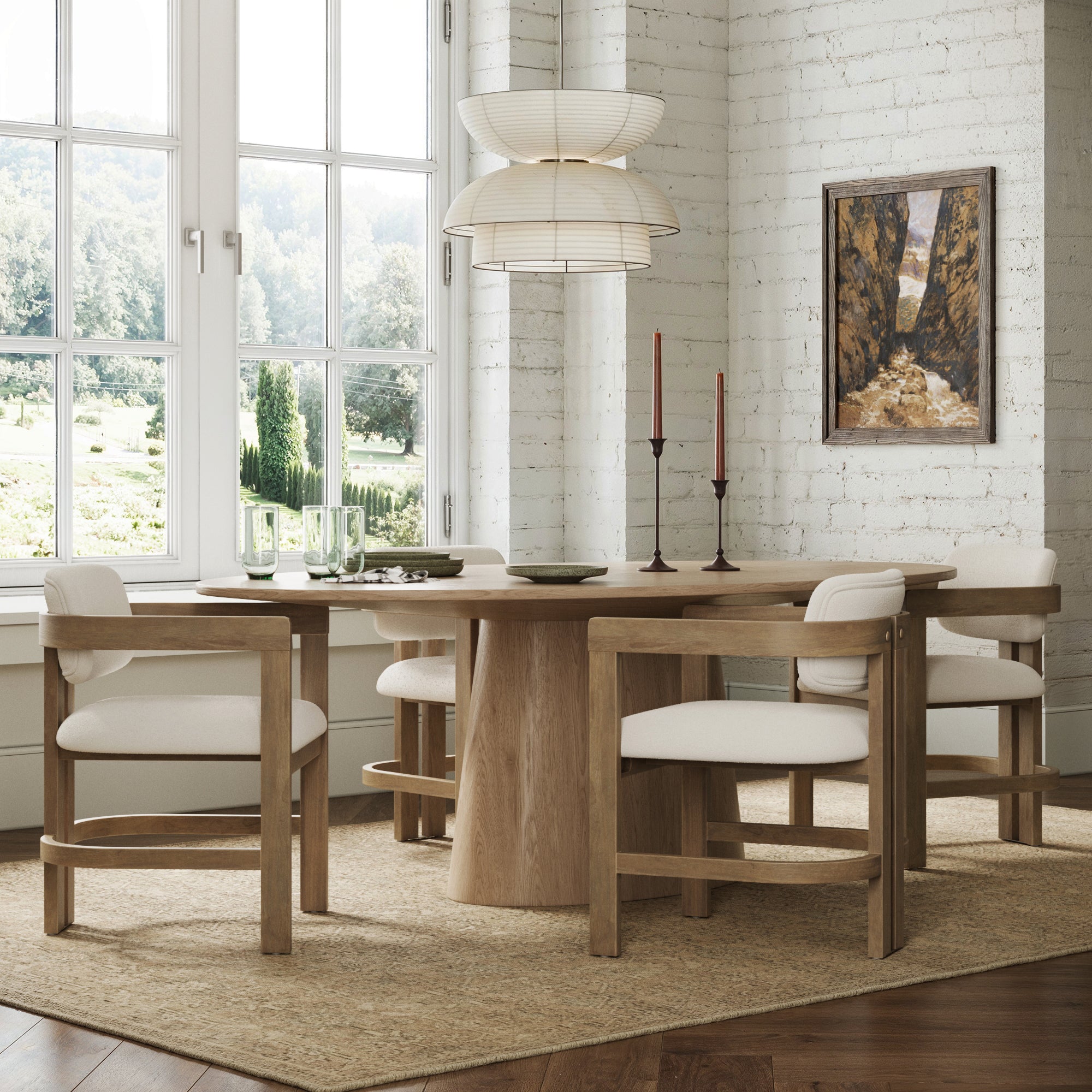 A modern dining room with a round wooden table, four Nathan James Boucle Dining Chairs (light brown), two tall red candles, glassware, a garden-view window, woven rug, pendant lamp, and framed painting on a white brick wall.