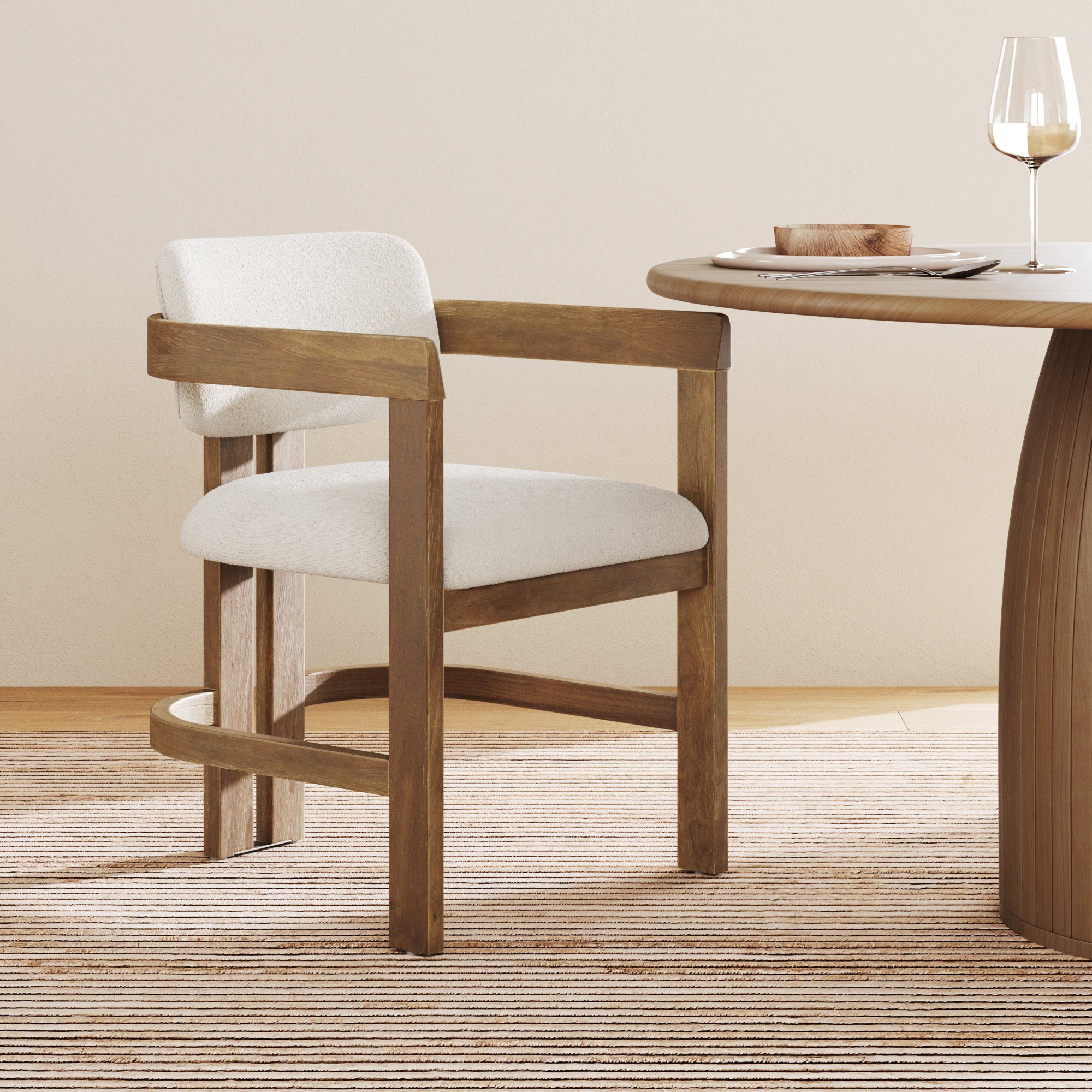 A Nathan James Boucle Dining Chair with a curved back and arms in light brown sits beside a round wooden dining table arranged with a wine glass, plate, and wooden bowl on a striped rug in a minimalist room.