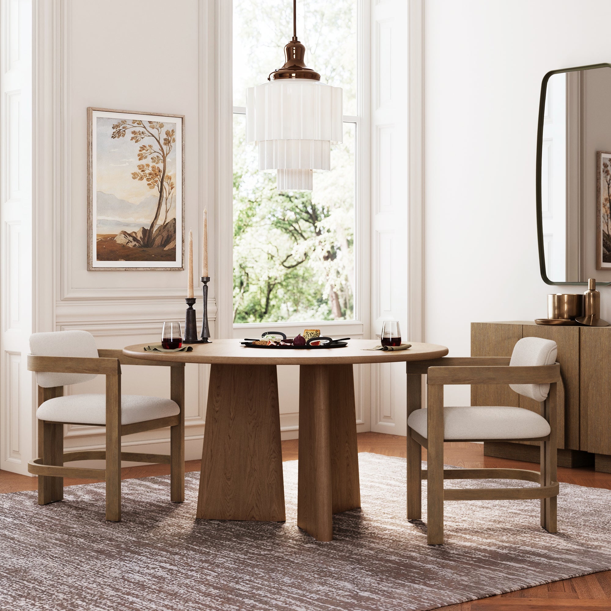 A modern dining room features a round wooden table, two Nathan James Boucle Dining Chairs with curved back and arm in light brown, candles, glassware on a tray, a large window, framed art, pendant light, sideboard, and a rug.