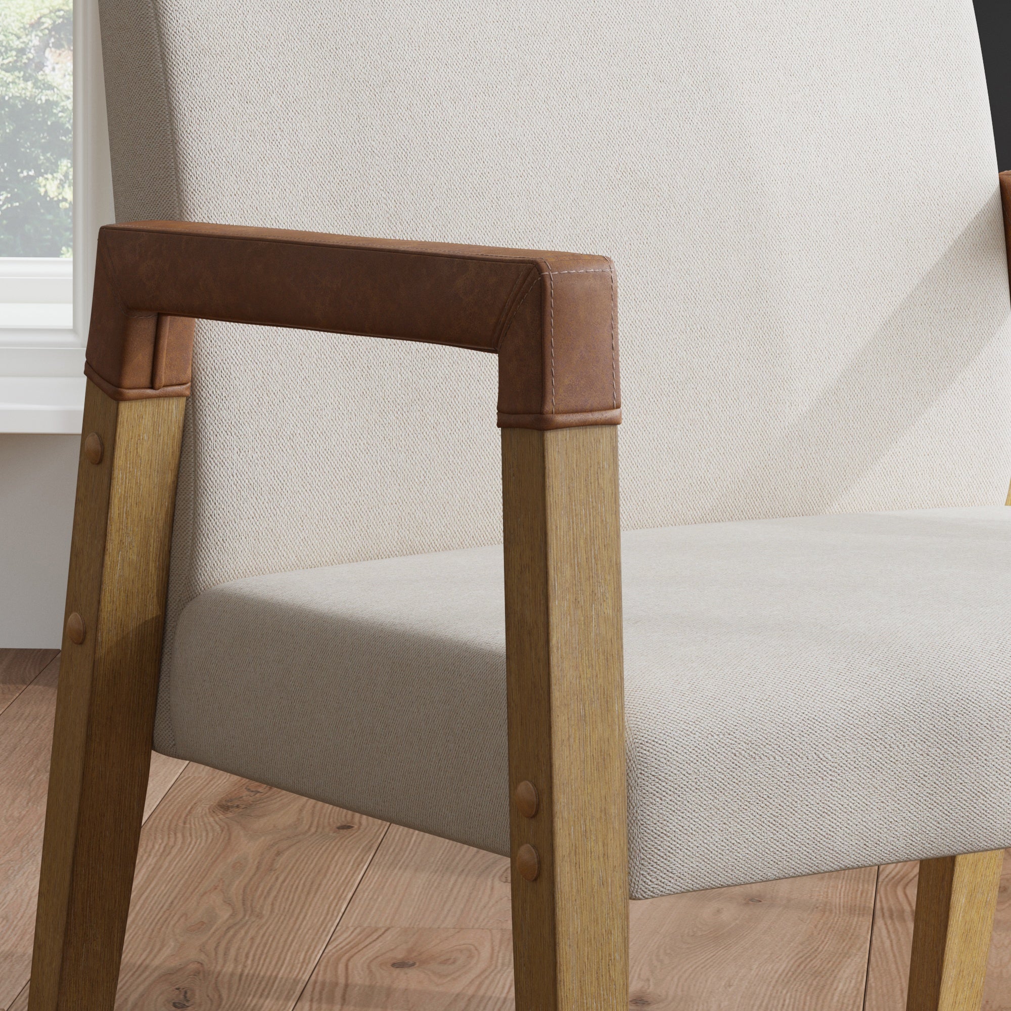 Close-up of a modern chair with light beige fabric upholstery, wooden legs, and brown leather-wrapped armrests, placed on a wooden floor near a window.