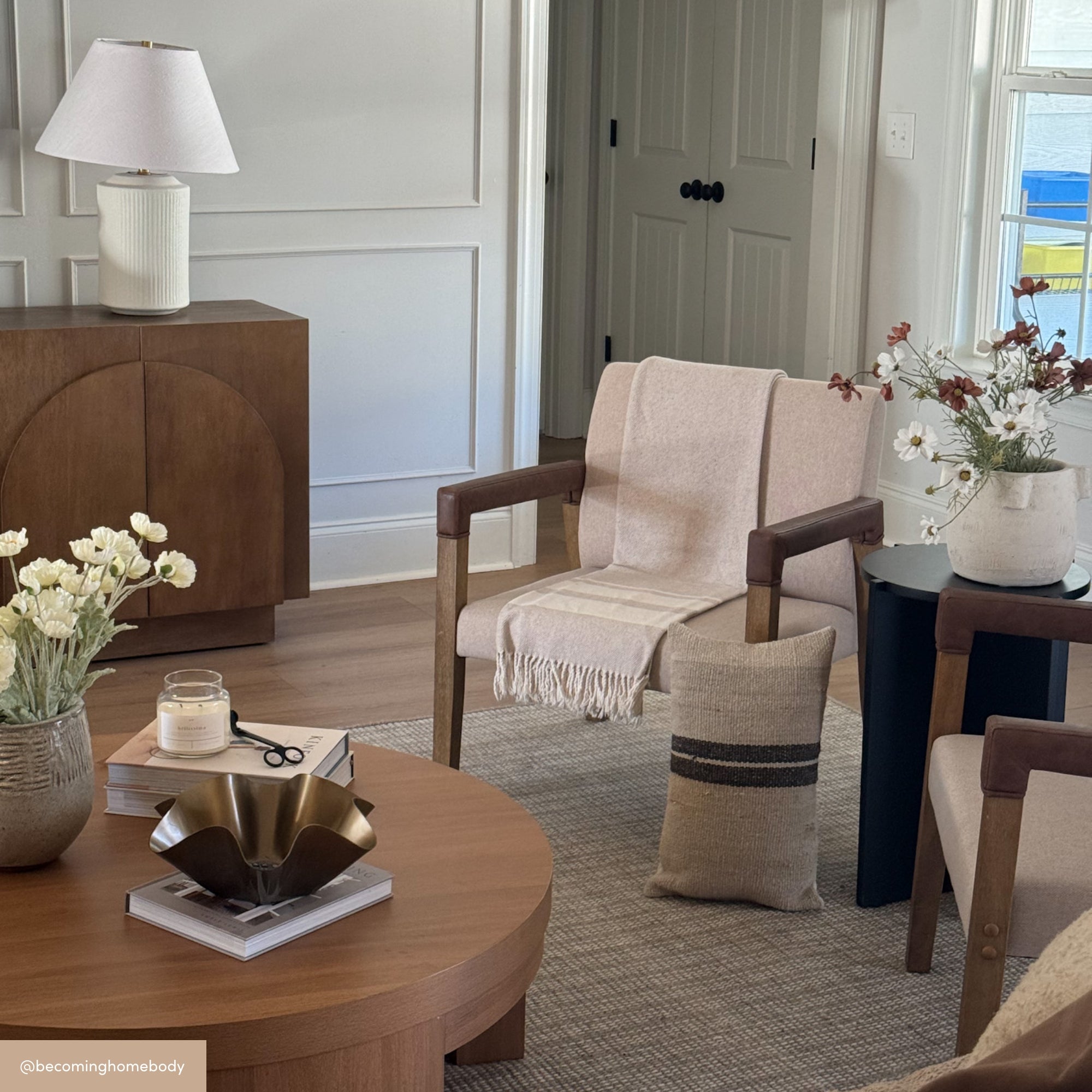 A cozy living room with wooden furniture, a round coffee table with decor, a beige blanket on a modern chair, neutral tones, a table lamp, and vases of dried flowers creating a warm, inviting atmosphere.