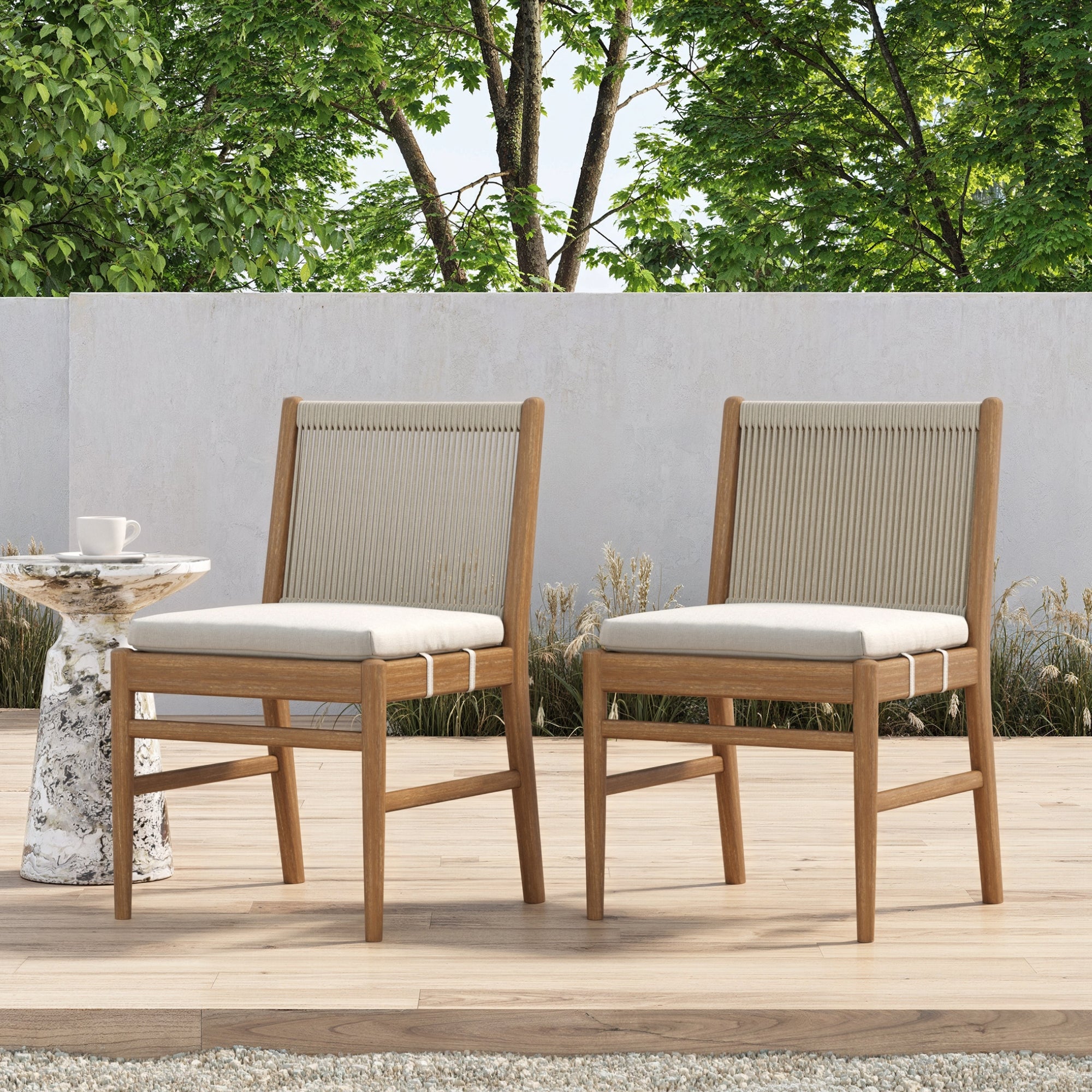 Two Nathan James Outdoor Wood & Rope Dining Chairs in brushed light brown are set on a wooden patio beside a small round side table with a cup, framed by lush green trees and a concrete wall for a cozy, serene ambiance.