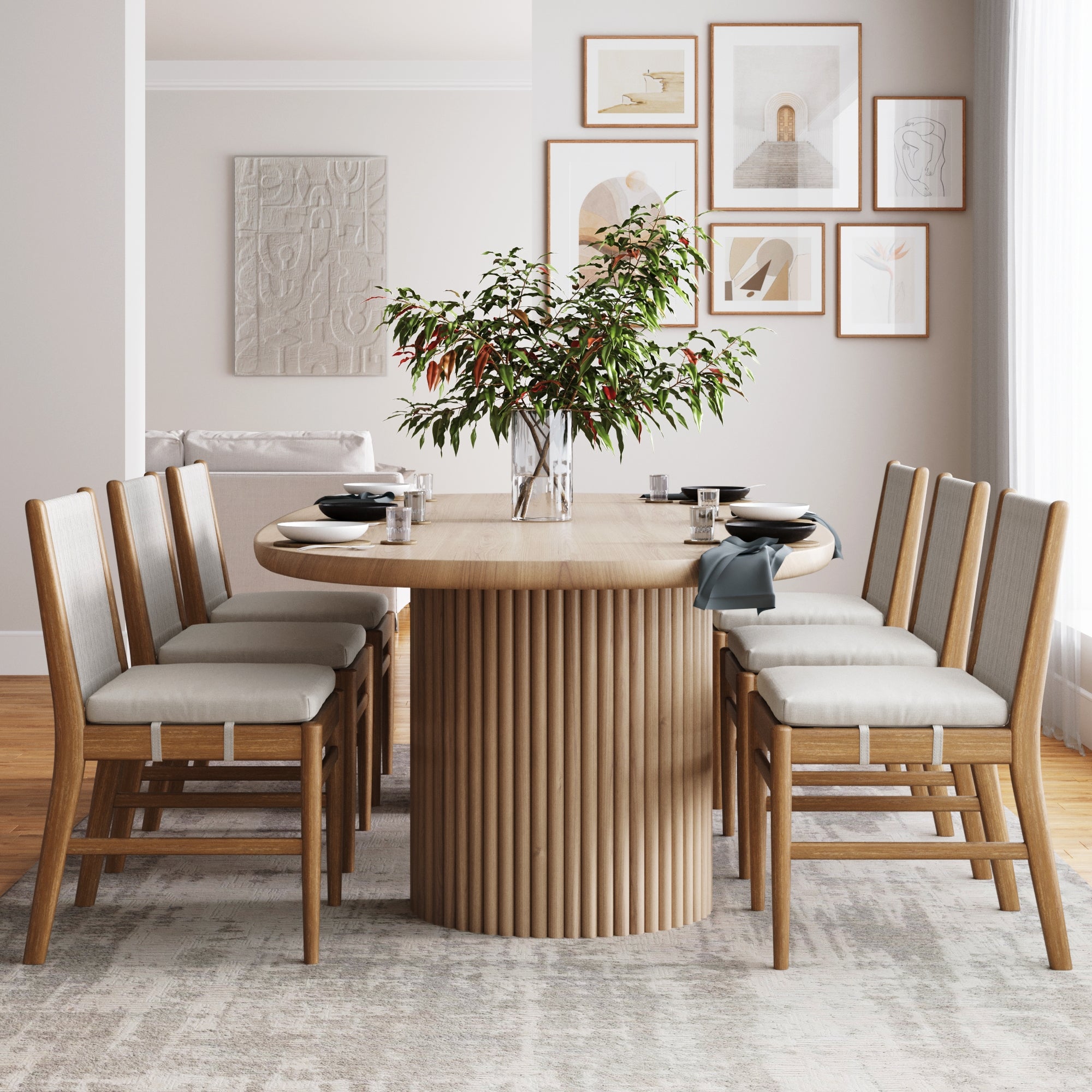 A modern dining room features a round wooden table, six Nathan James Woven Rope Back Dining Chairs in brushed light brown with upholstered seats, a leafy branch centerpiece, framed art on pale walls, and sunlight streaming through white curtains.
