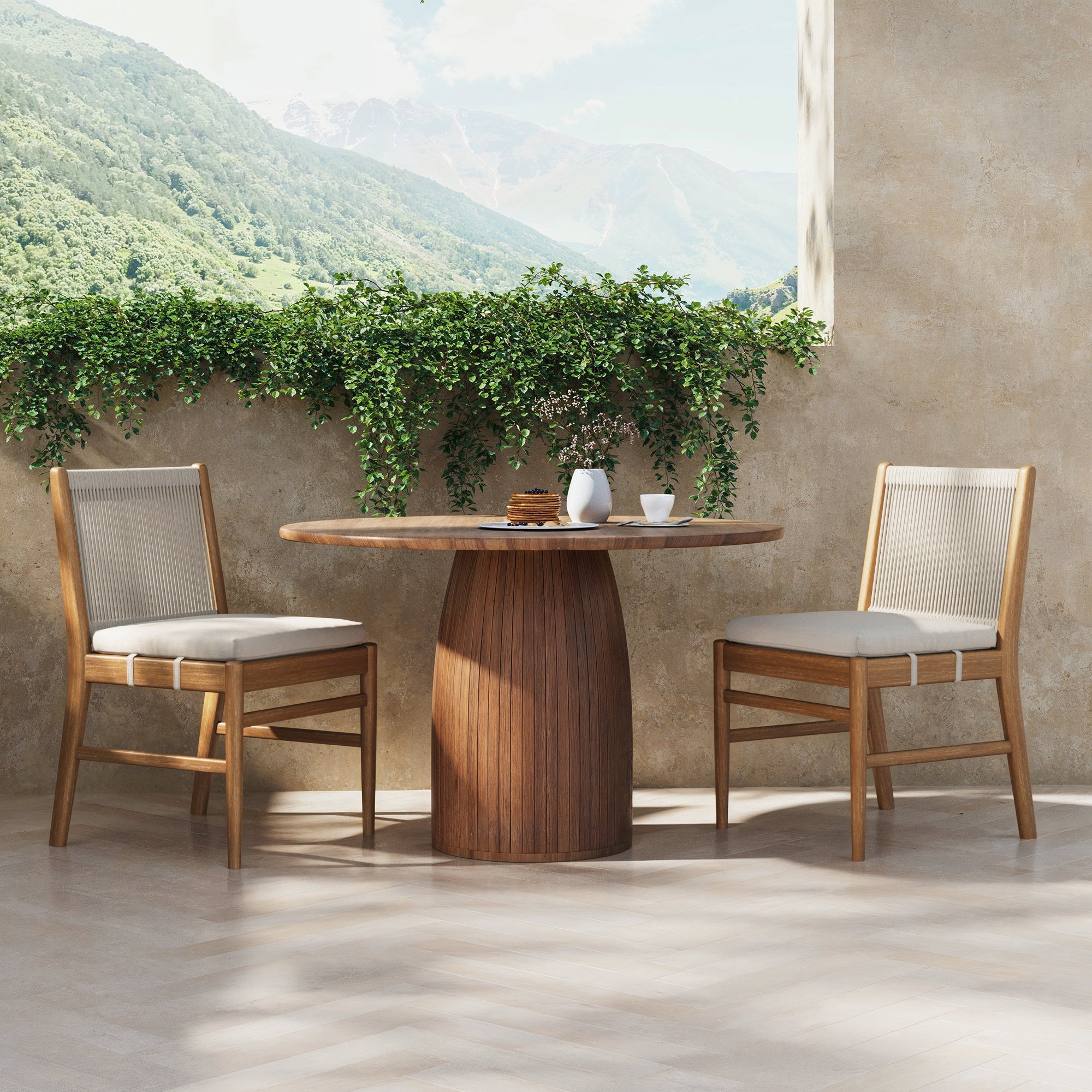 A round wooden table with two Nathan James Outdoor Wood & Rope Dining Chairs in brushed light brown sits by a balcony with mountain views and green plants. A coffee cup, teapot, and pastries on the table create a cozy morning atmosphere.