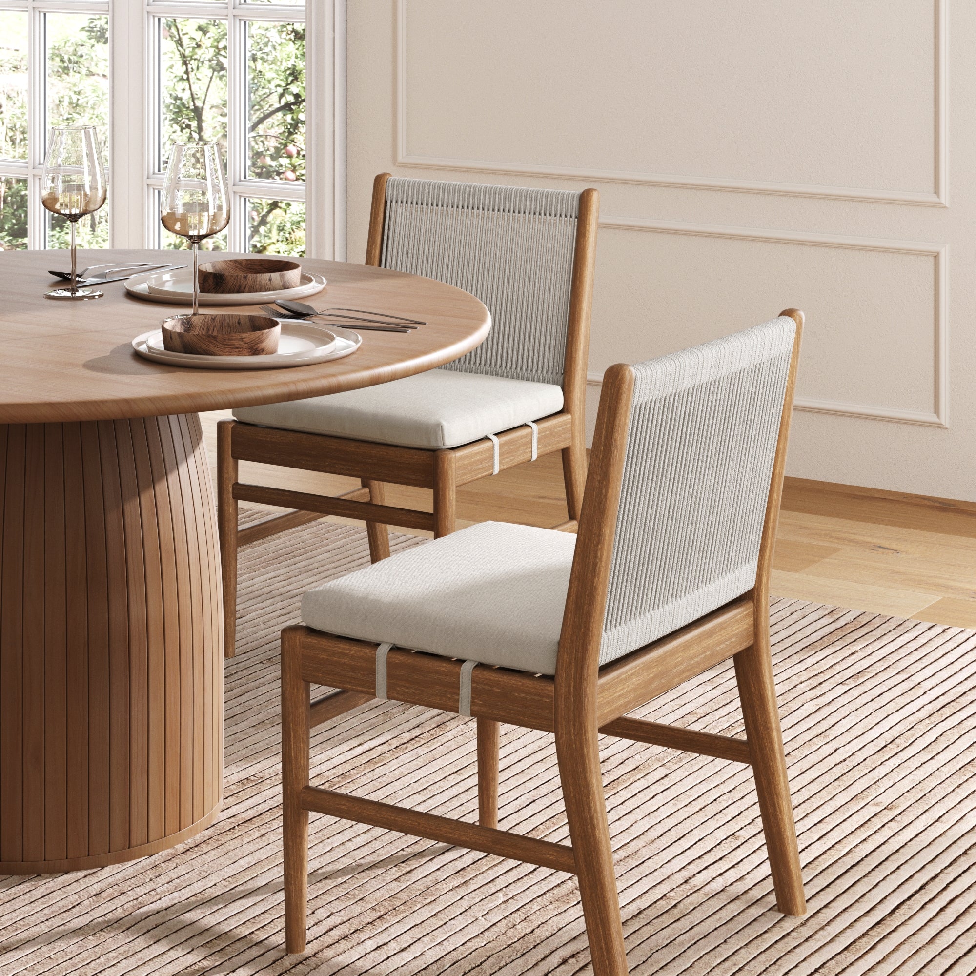 A cozy dining area with a round wooden table set for two features Nathan James Woven Rope Back Dining Chairs in brushed light brown, their solid wood frames and woven rope backrests resting on a striped rug with sunlight streaming through the window.