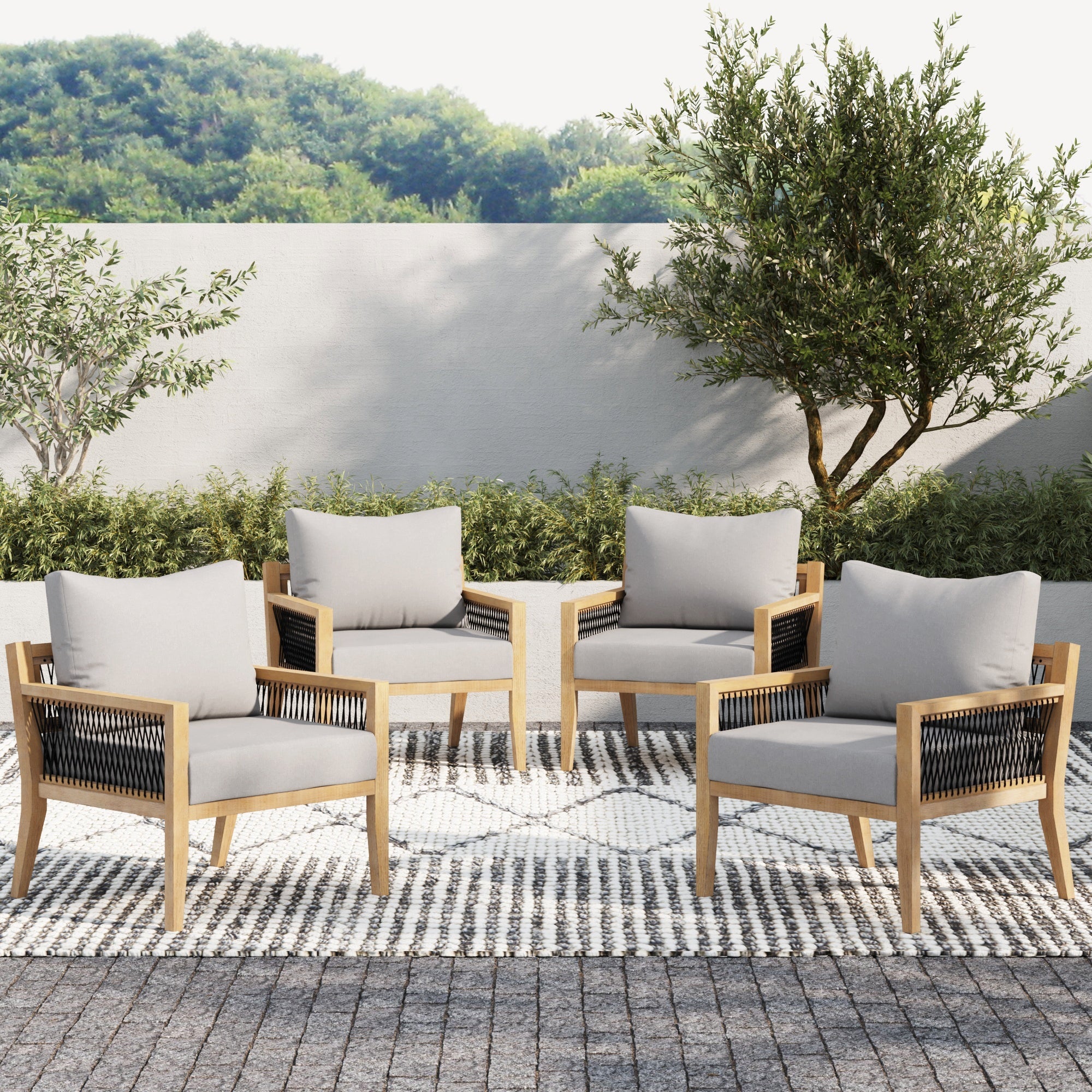 Four Nathan James Outdoor Wood Patio Arm Chairs in gray acacia with removable cushions surround a small side table on a patterned rug, set against lush greenery and a white wall.