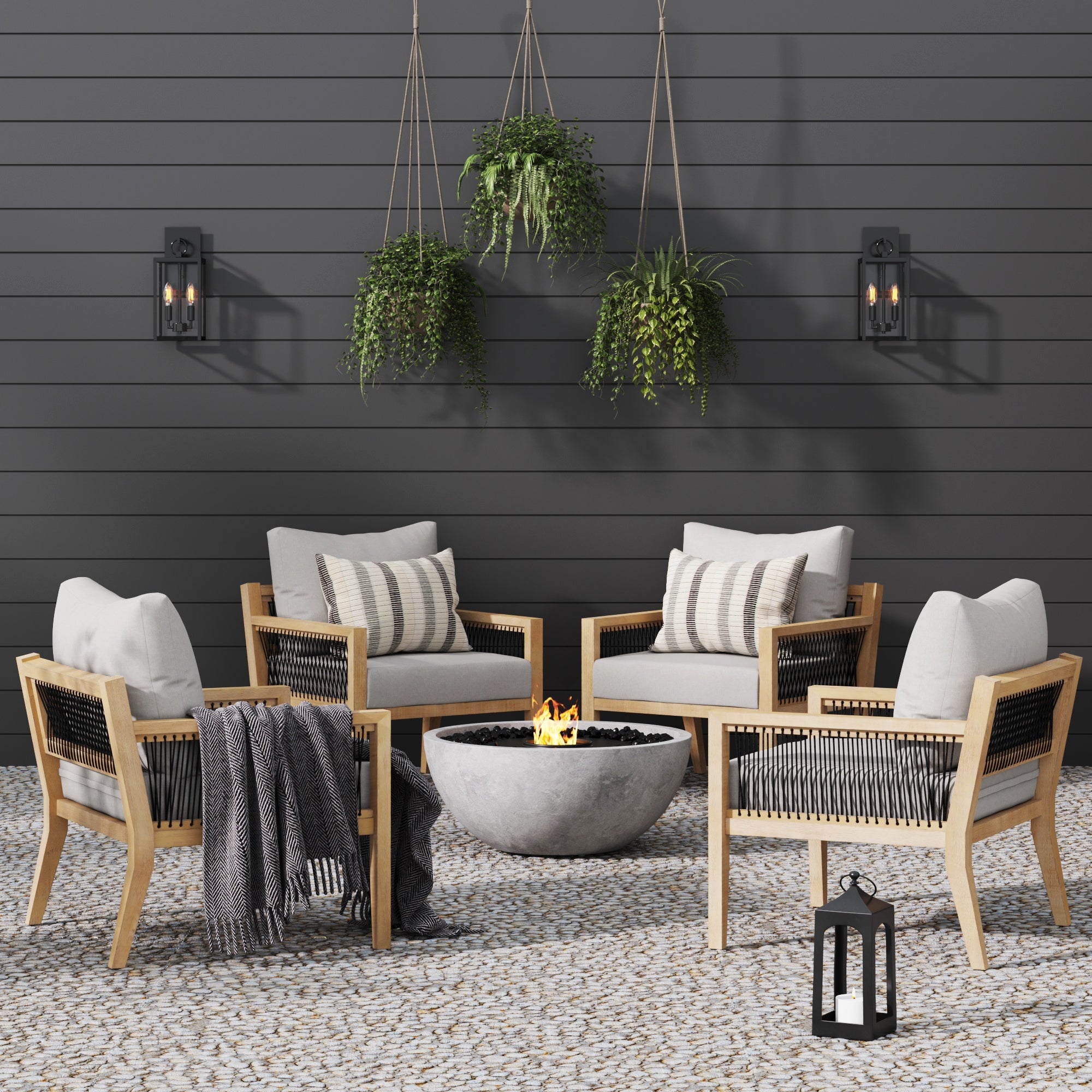 Four cushioned wooden patio chairs surround a modern round fire pit on a pebble patio, with a lantern, throw blanket, hanging plants, and wall-mounted lights against a dark horizontal wood wall.