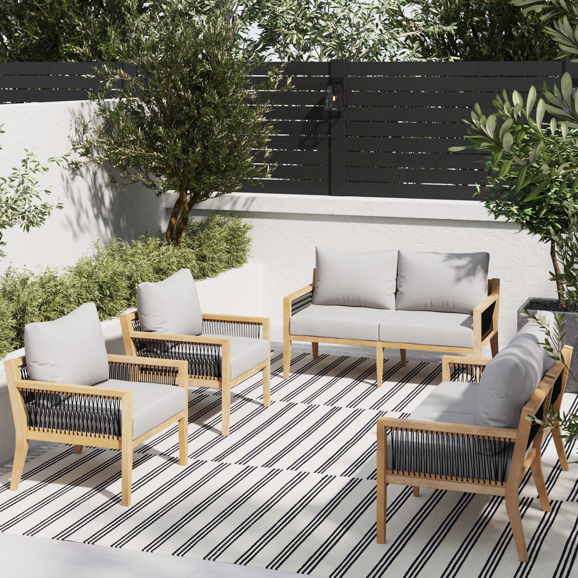 Outdoor patio with four wooden-framed chairs and a loveseat with gray cushions, arranged on a striped rug. Surrounded by green shrubs, trees, and a black privacy fence.