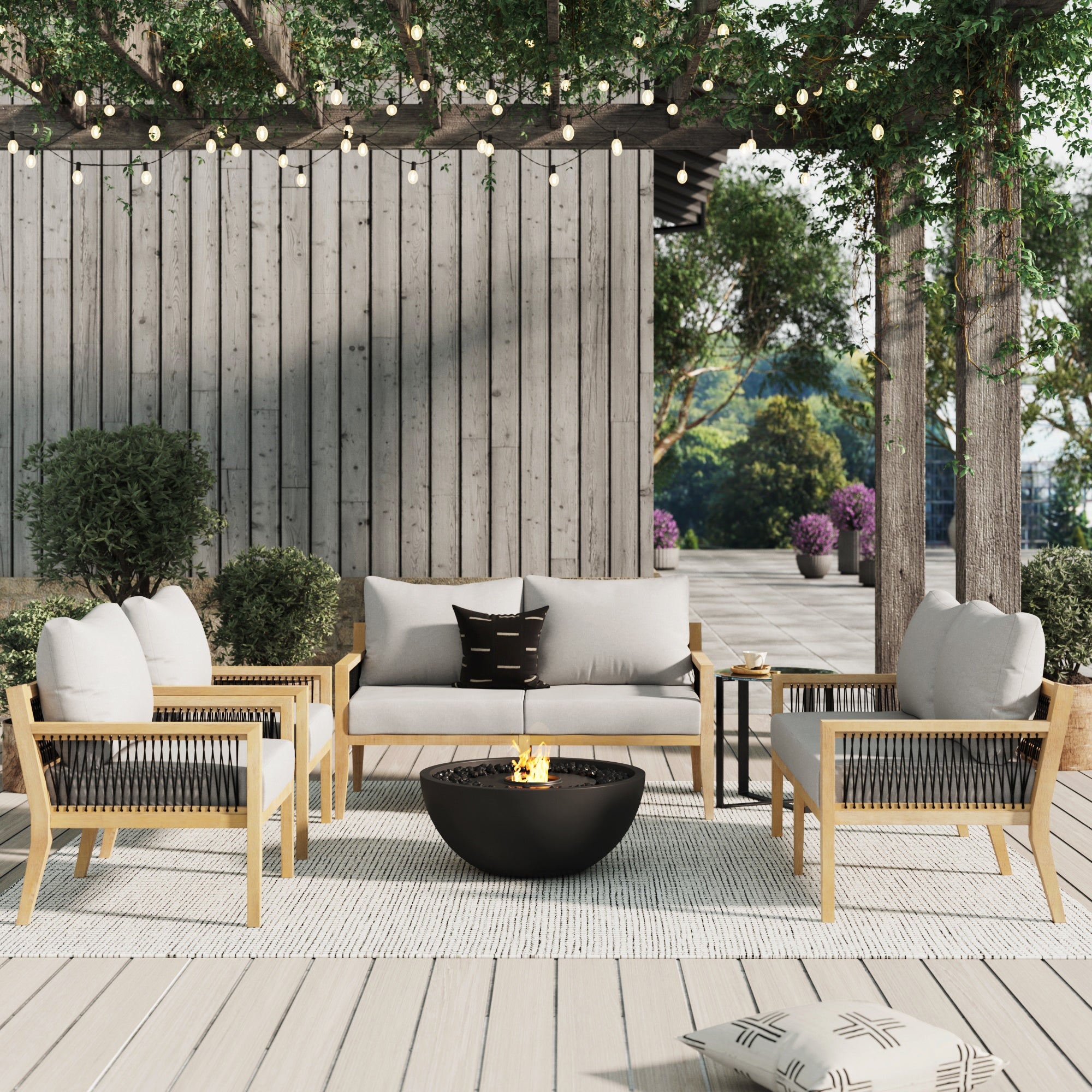 Modern outdoor patio with a gray sofa, two armchairs, a round black fire pit, and string lights overhead. The setting includes trees, greenery, and a wooden privacy fence in the background.