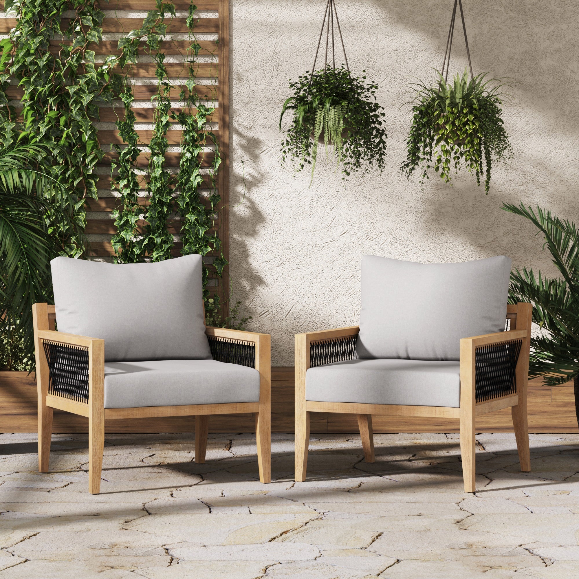 Two modern wooden lounge chairs with light gray cushions are placed on a stone patio, surrounded by lush green plants and hanging planters against a beige wall with a wooden trellis.