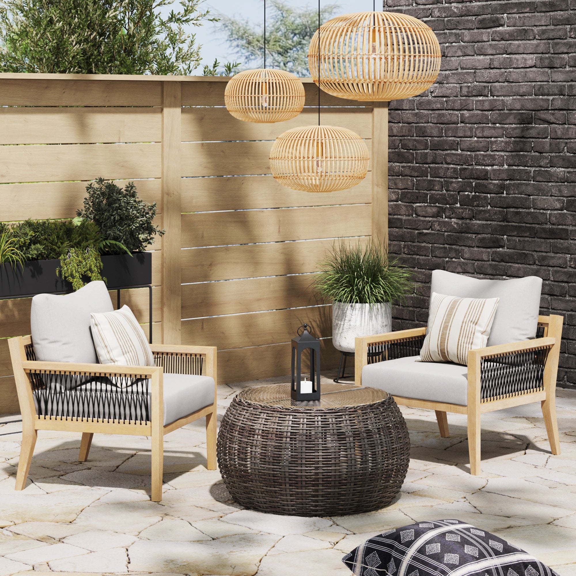 Two Nathan James Outdoor Wood Patio Arm Chairs in gray acacia, each with removable cushion covers and striped pillows, sit around a round wicker coffee table with a lantern. Three rattan pendant lights hang overhead amid lush plants and textured flooring.
