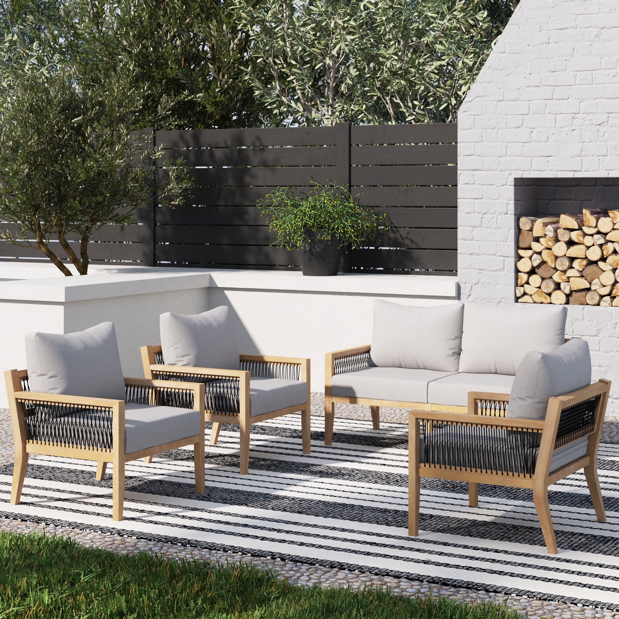 Outdoor patio seating area with a striped rug, two armchairs, a loveseat, and a coffee table featuring wood frames and light gray cushions. A white brick fireplace with stacked firewood is in the background.