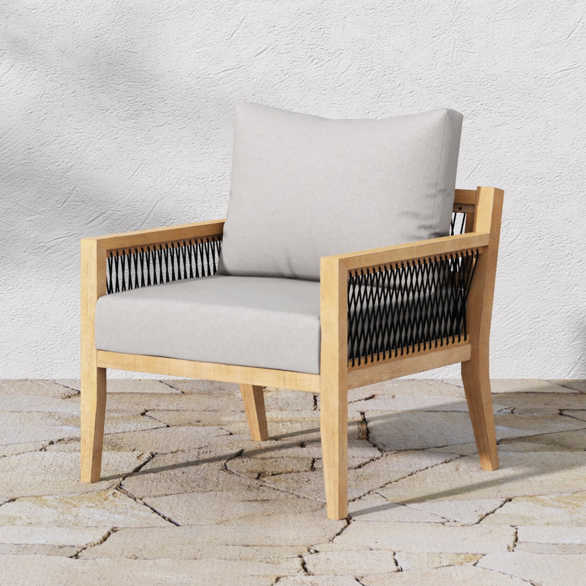 The Nathan James Outdoor Patio Wood Arm Chair, made of weatherproof gray acacia with beige removable cushions and black woven sides, sits on a stone patio against a light textured wall.