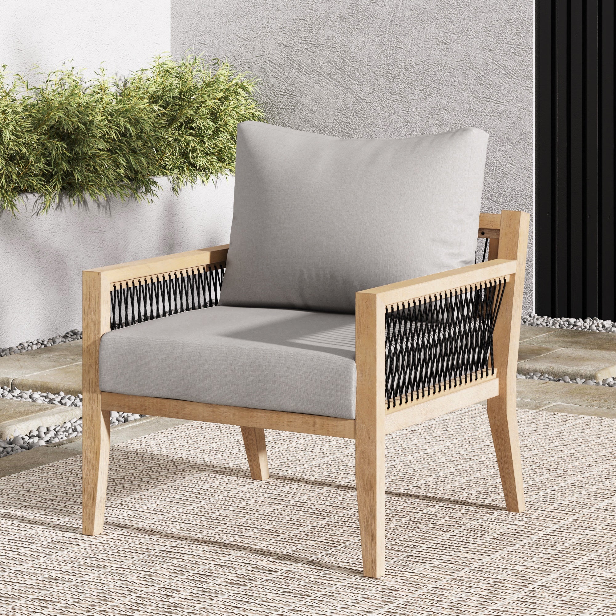A modern outdoor armchair with light wood frame, light gray cushioned seat and back, and black woven sides, sits on a textured beige rug. Green plants and a white wall are in the background.