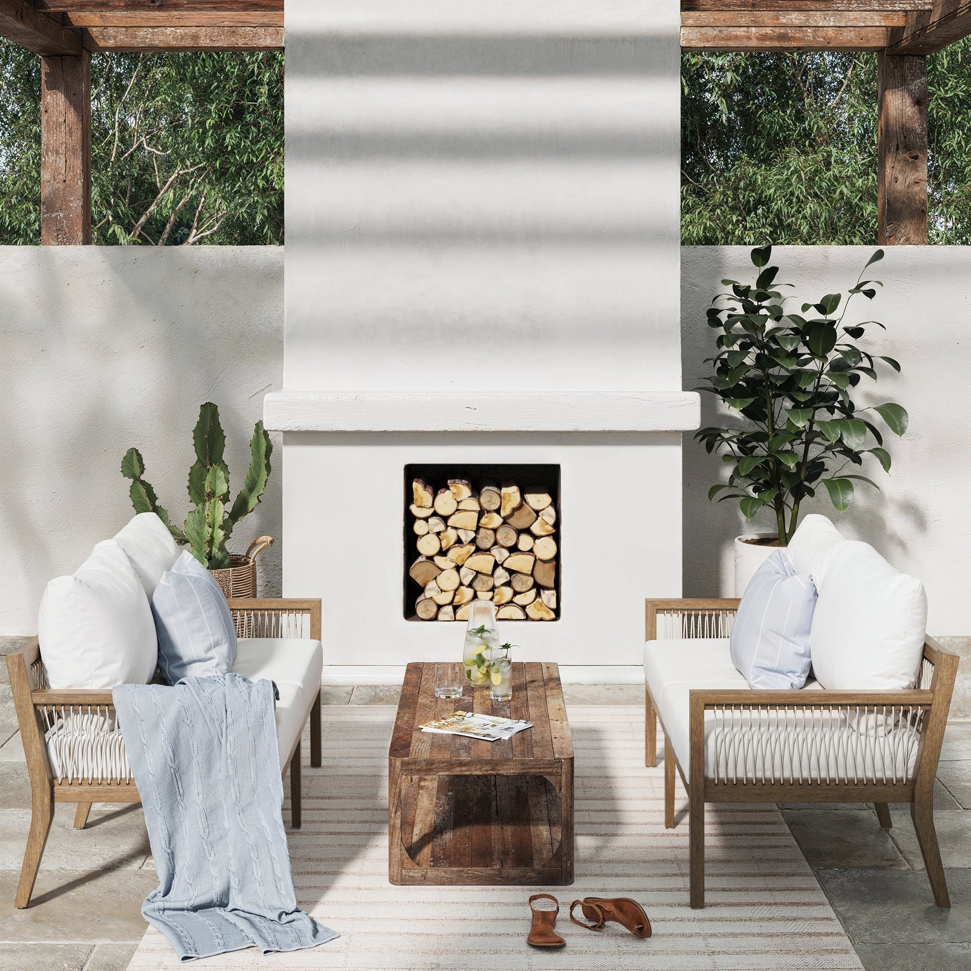 The Nathan James Outdoor Patio Wood Set includes 2 light brown loveseats with white, UV fade-resistant cushions, a coffee table, and cozy accents like a blue throw, potted plants, sandals, and a striped rug for a stylish outdoor retreat.