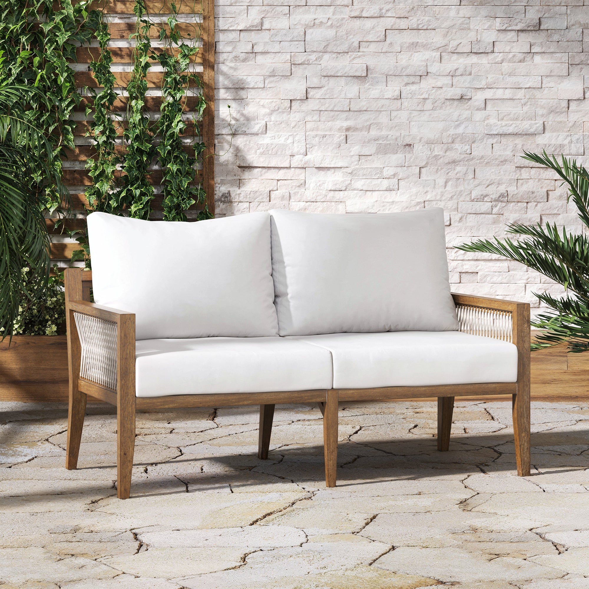 A wooden loveseat with white cushions sits on a stone patio. Green plants and vines climb a trellis and a white stone wall in the background, creating a cozy outdoor setting.