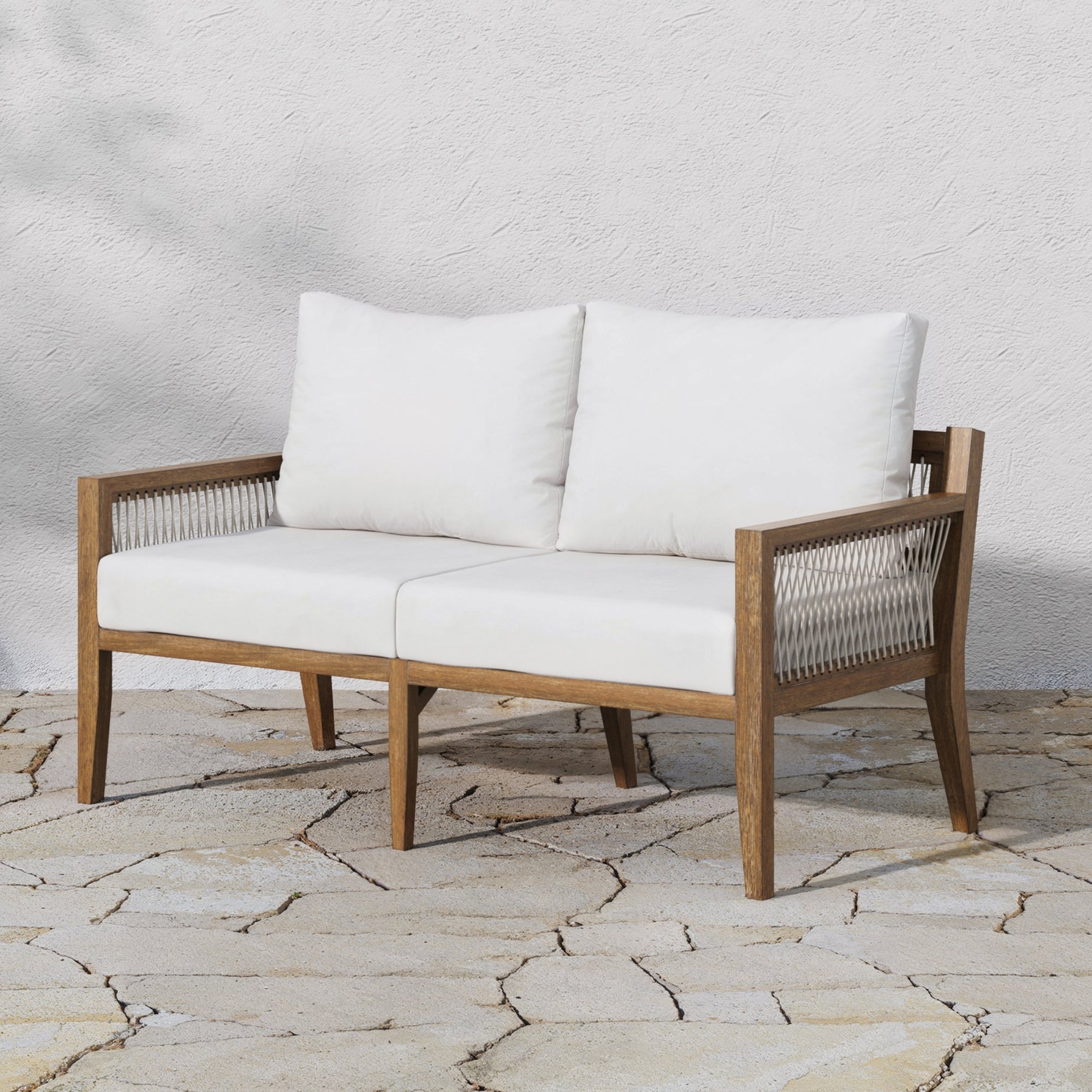 The Nathan James Outdoor Wood Patio Loveseat in white and light brown features weather-resistant woven rope sides and plush white cushions, creating stylish outdoor seating against a textured white wall on a stone patio.