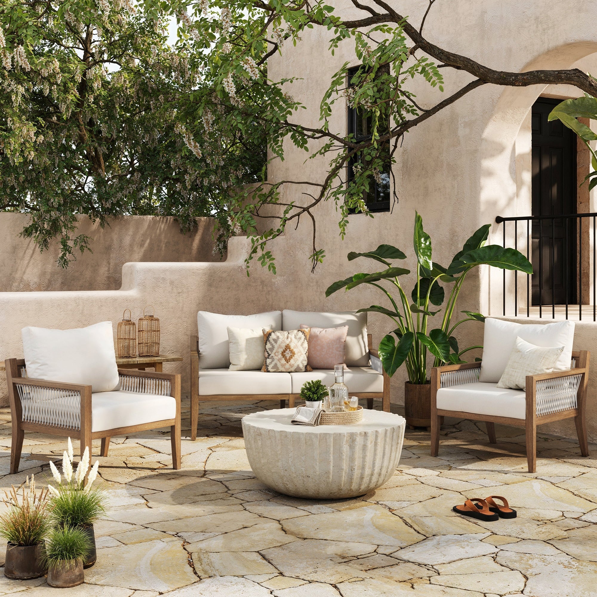 Outdoor patio with stone flooring, wooden furniture with white cushions, a round stone coffee table, potted plants, and a tree overhead. Beige stucco building and steps in the background create a Mediterranean vibe.