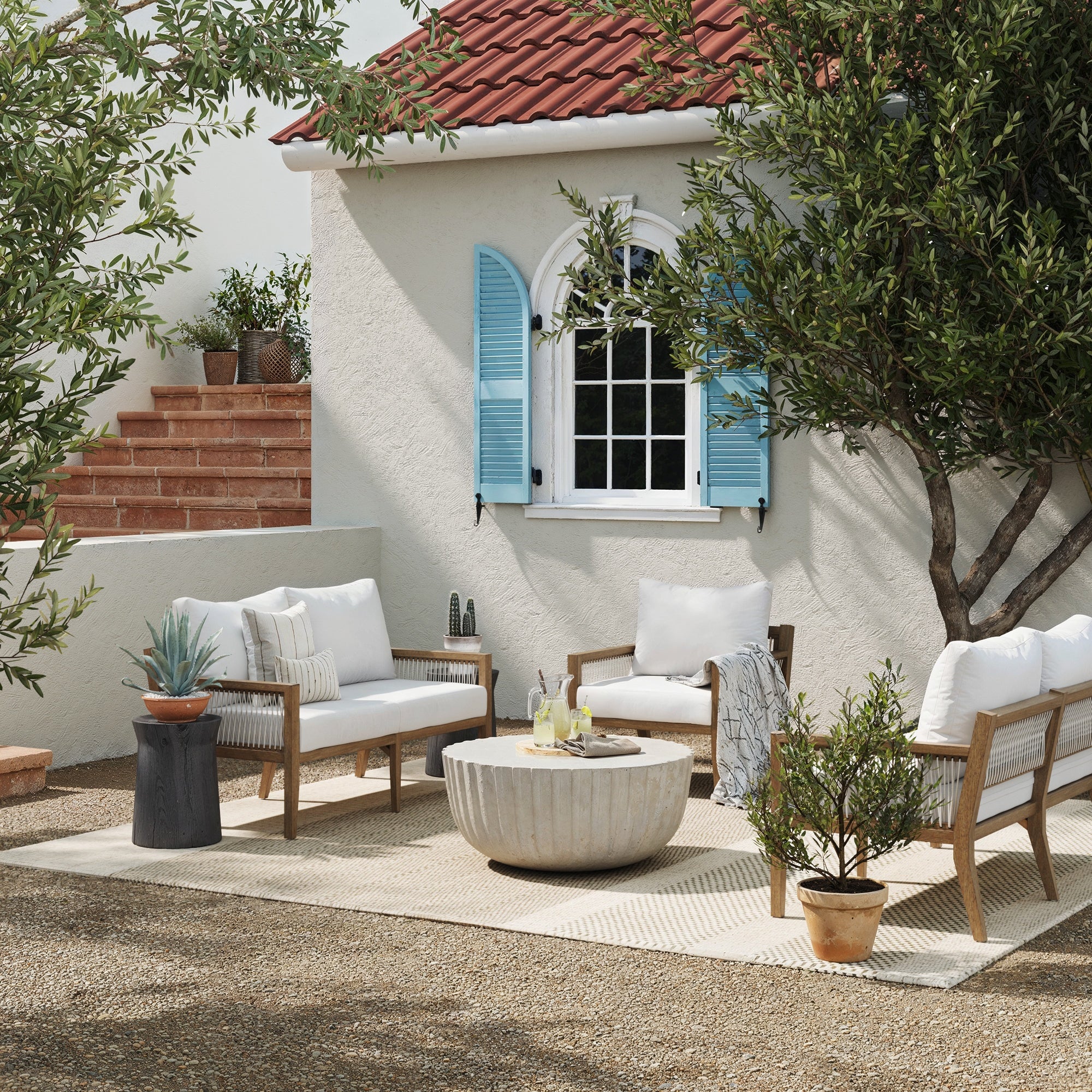 Outdoor patio scene with white cushioned wooden furniture, a round stone coffee table, potted plants, and a white stucco house with blue shutters and a red tile roof in the background.