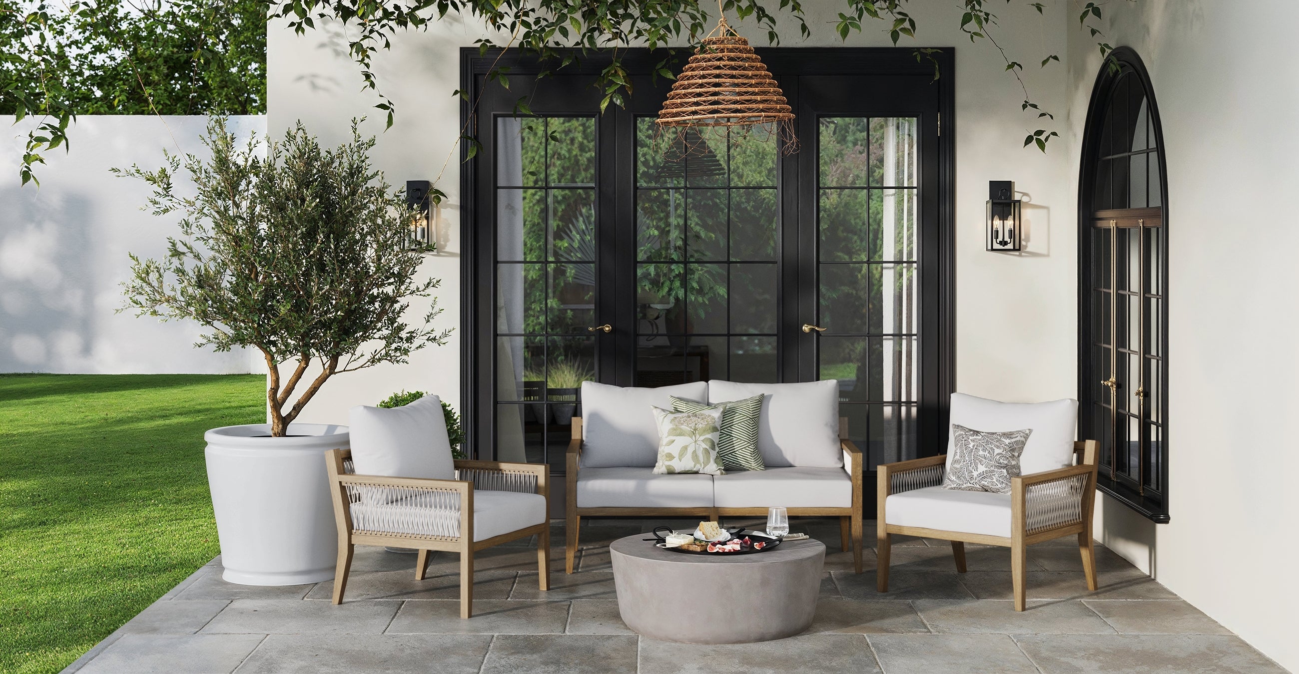 Outdoor patio with light wooden furniture and white cushions, round concrete table with snacks, potted tree, black-framed glass doors, wall lights, and wicker pendant lamp against a cream-colored wall. Lush green lawn in the background.