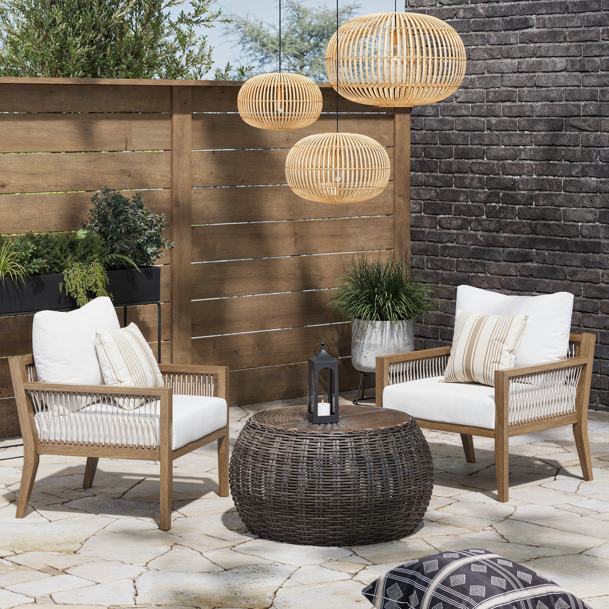 A cozy patio features a set of 2 Nathan James Outdoor Patio Wood Arm Chairs with white cushions, a round wicker coffee table, potted plants, and three hanging wicker lanterns against a wooden fence and brick wall.