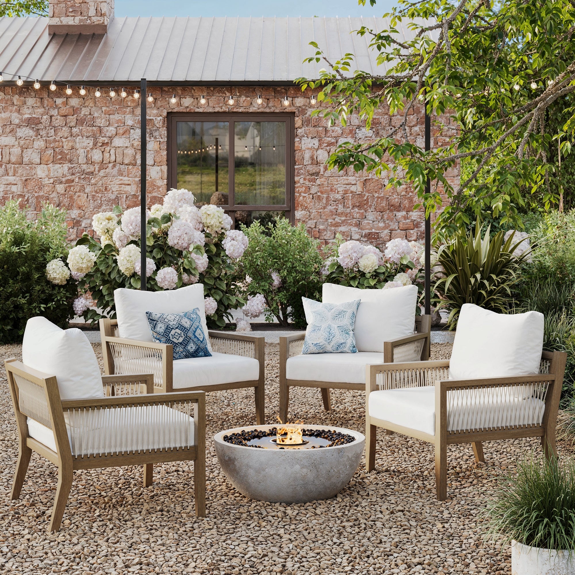 Four Nathan James Outdoor Patio Wood Arm Chairs in white are arranged around a round fire pit with decorative pillows, set on gravel amidst blooming hydrangeas, string lights, and a stone building for a perfect bohemian garden setting.