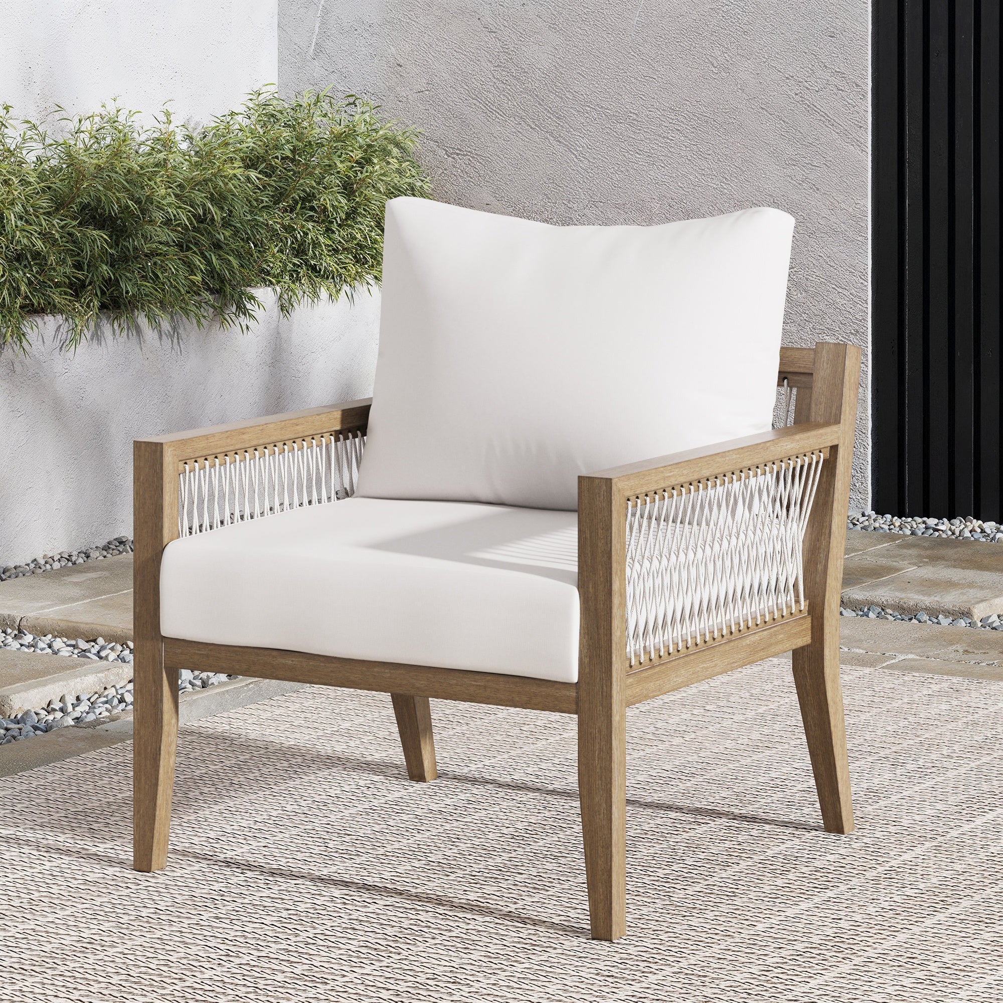 A modern outdoor armchair with a wooden frame, white seat and back cushions, and woven side panels, placed on a textured rug with plants and a stone floor in the background.