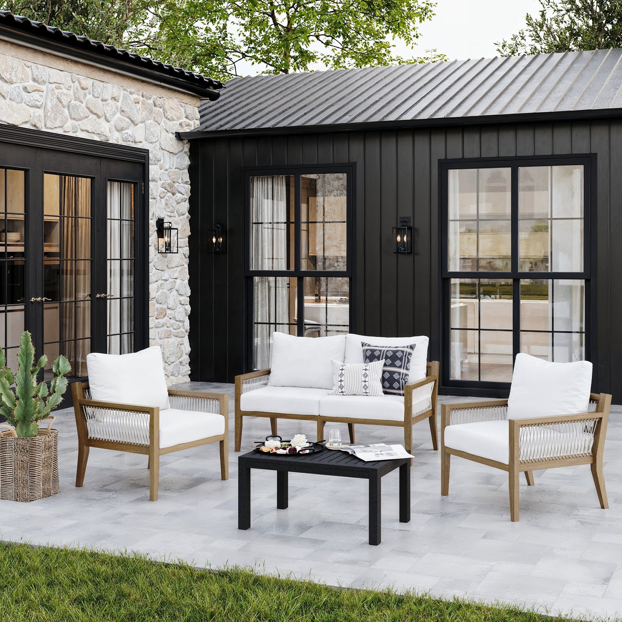 Modern outdoor patio with a black house exterior, stone wall, large windows, and a seating area featuring wooden chairs, a loveseat with white cushions, a black coffee table, and a potted cactus on tiled flooring.