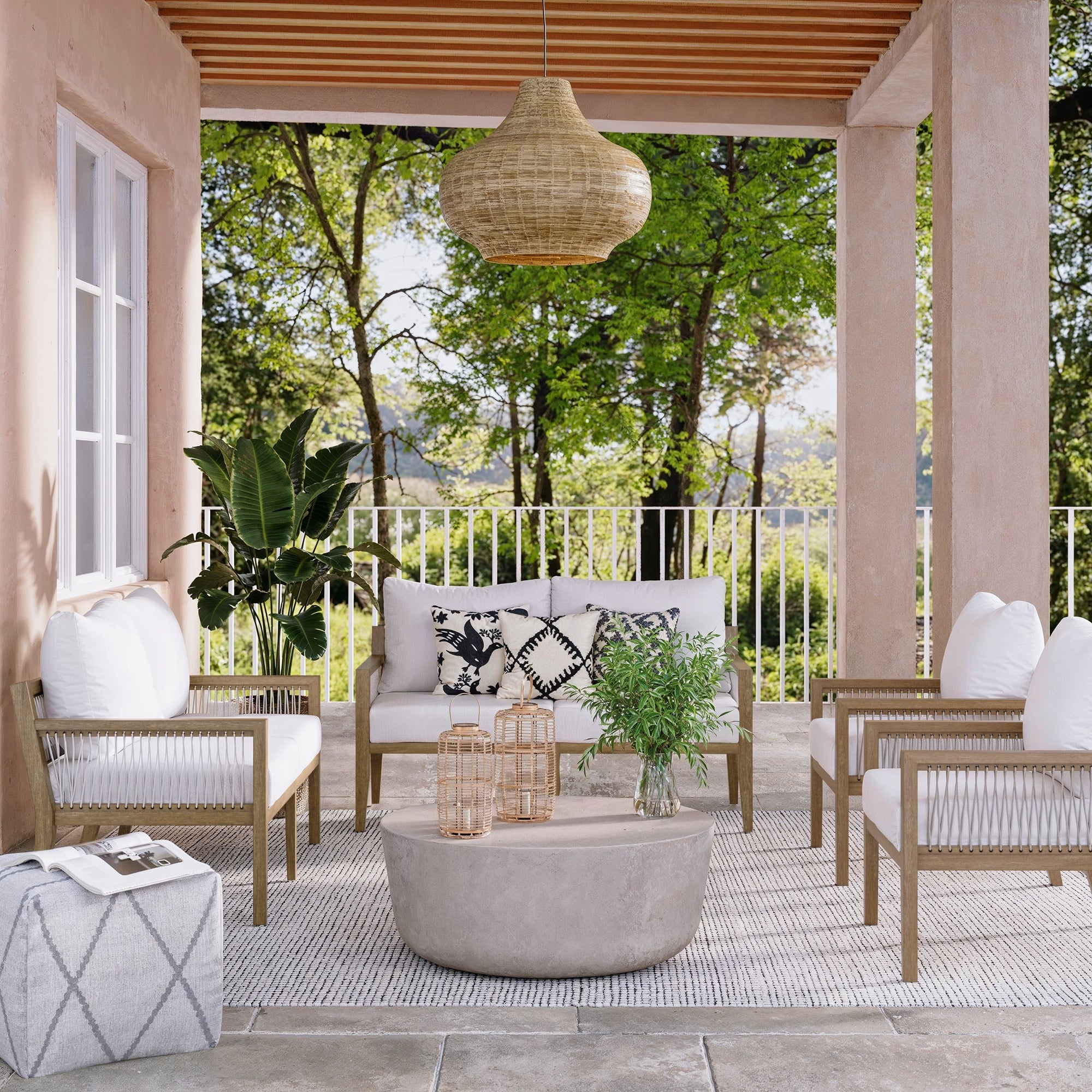 A stylish outdoor patio with a sofa, two armchairs, a round coffee table, and pouf on a striped rug. Cushions and patterned pillows decorate the seating. Plants and lanterns add accents; trees and greenery are visible in the background.