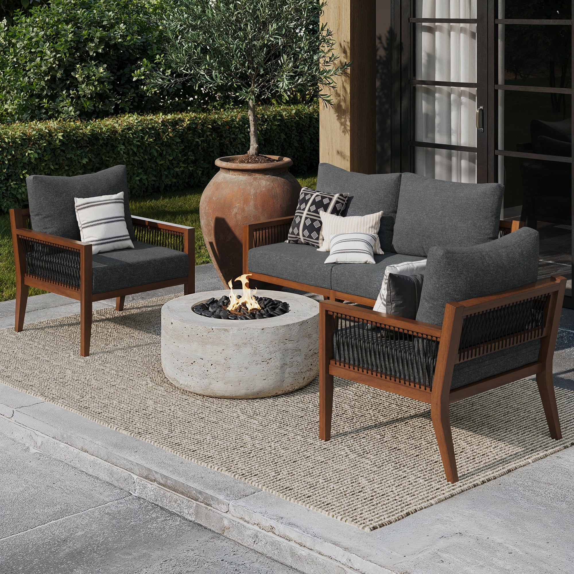 An outdoor patio features the Nathan James Outdoor 3 Piece Wood Patio Set with a loveseat and chairs, all with gray cushions, arranged around a round concrete fire pit on a beige rug beside a house and ceramic planter, surrounded by greenery.
