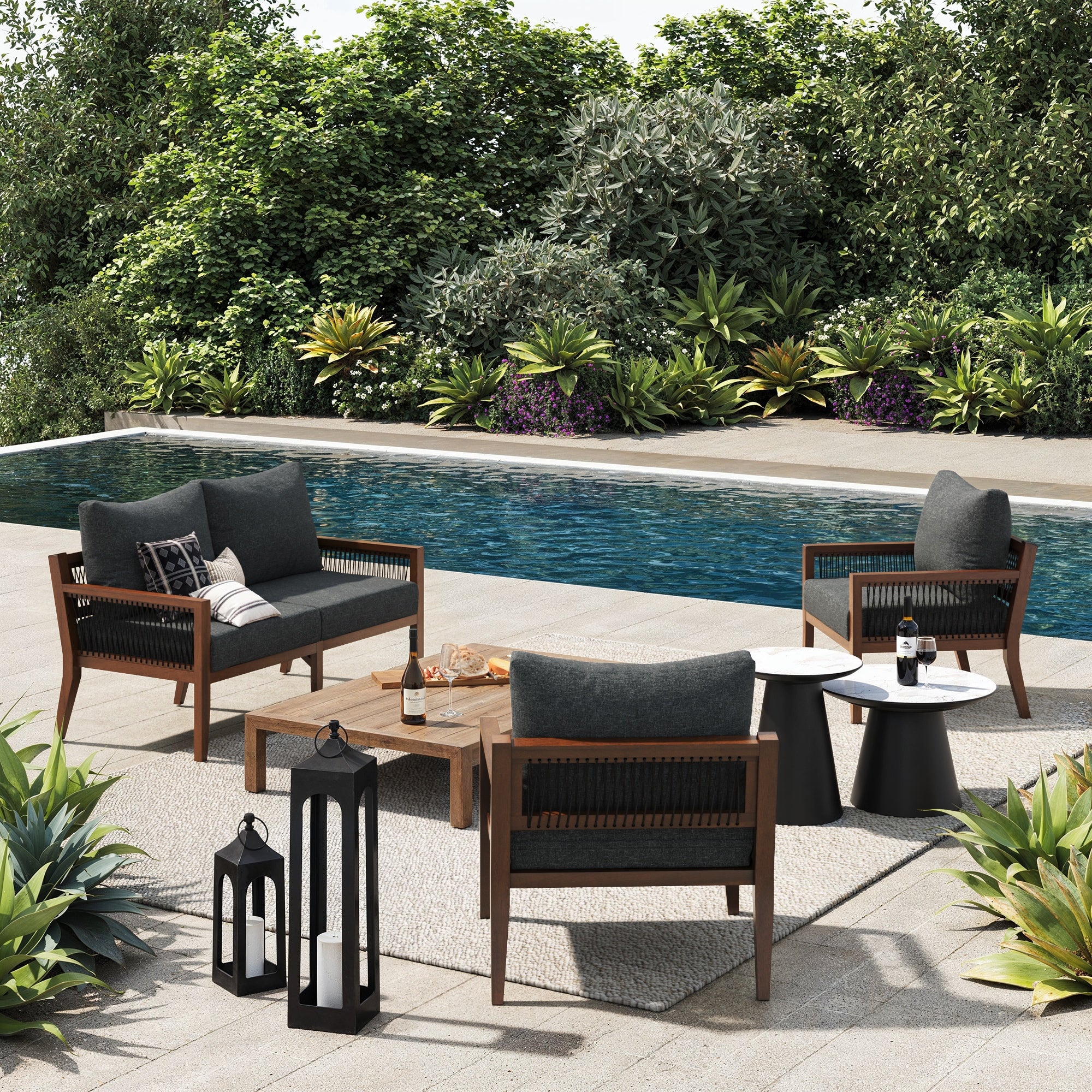 Relax by the pool with the Nathan James Outdoor 3 Piece Wood Patio Set Loveseat and Chairs, featuring dark cushioned seating, a wooden table, lanterns, side tables, and wine. Lush green plants surround this inviting oasis.