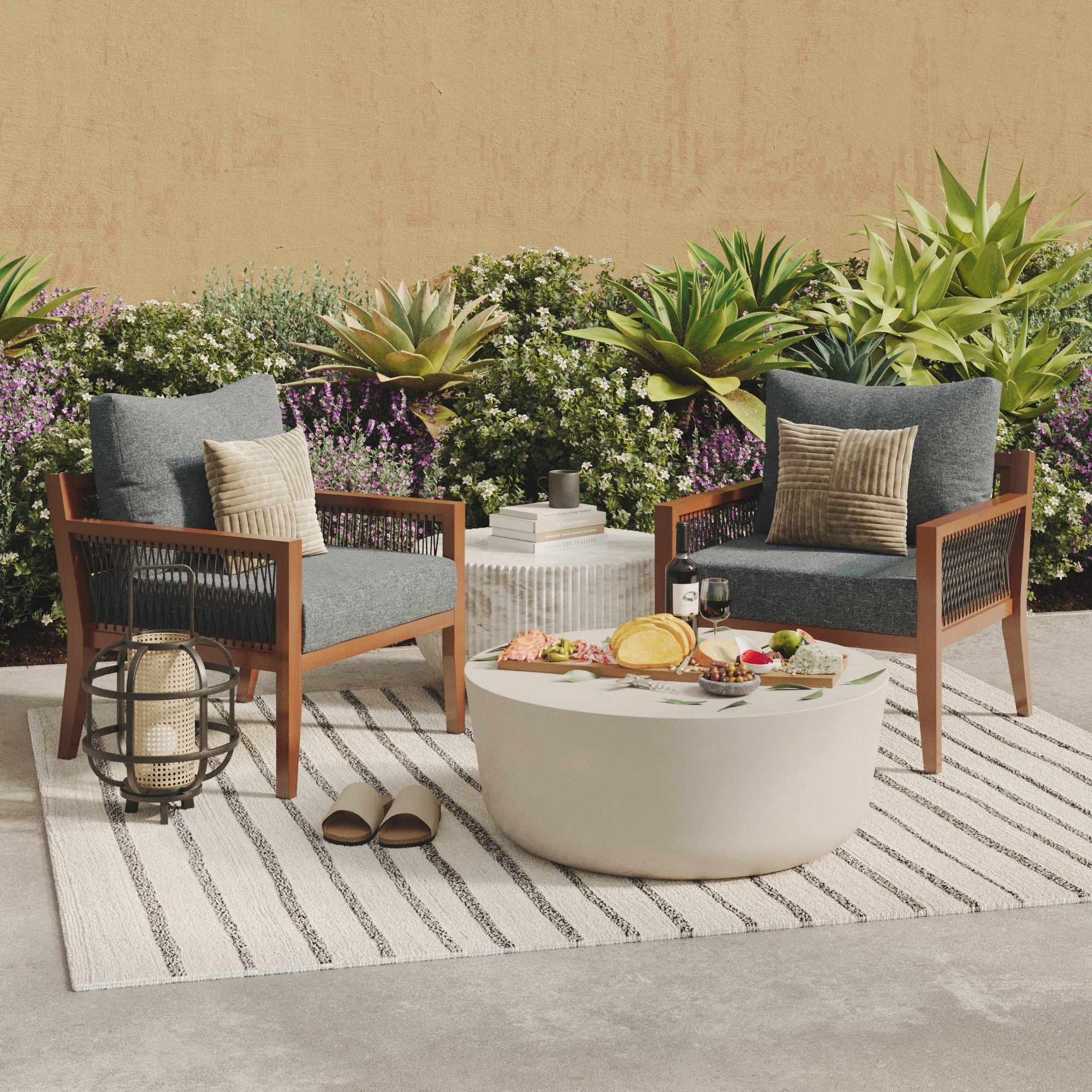 Two cushioned wooden chairs with pillows sit on a striped rug beside a round table with food and drinks. The setup is outdoors, decorated with plants, slippers, and a lantern, against a beige wall and green shrubbery.