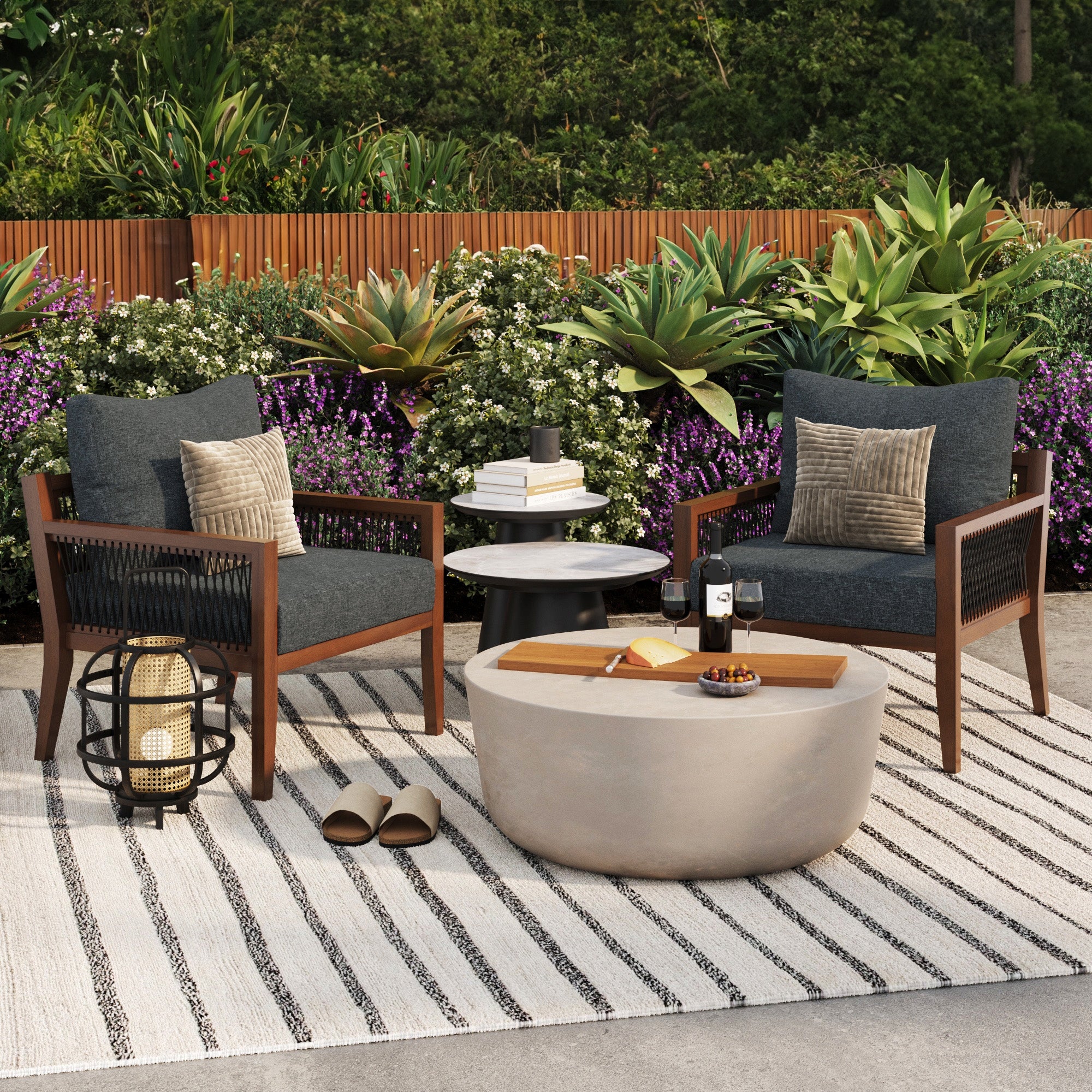 Two Nathan James Outdoor Patio Wood Arm Chairs in gray with washable cushions sit on a striped rug, facing a concrete coffee table with wine, cheese, and books. Plants and flowers complete the cozy garden patio atmosphere.