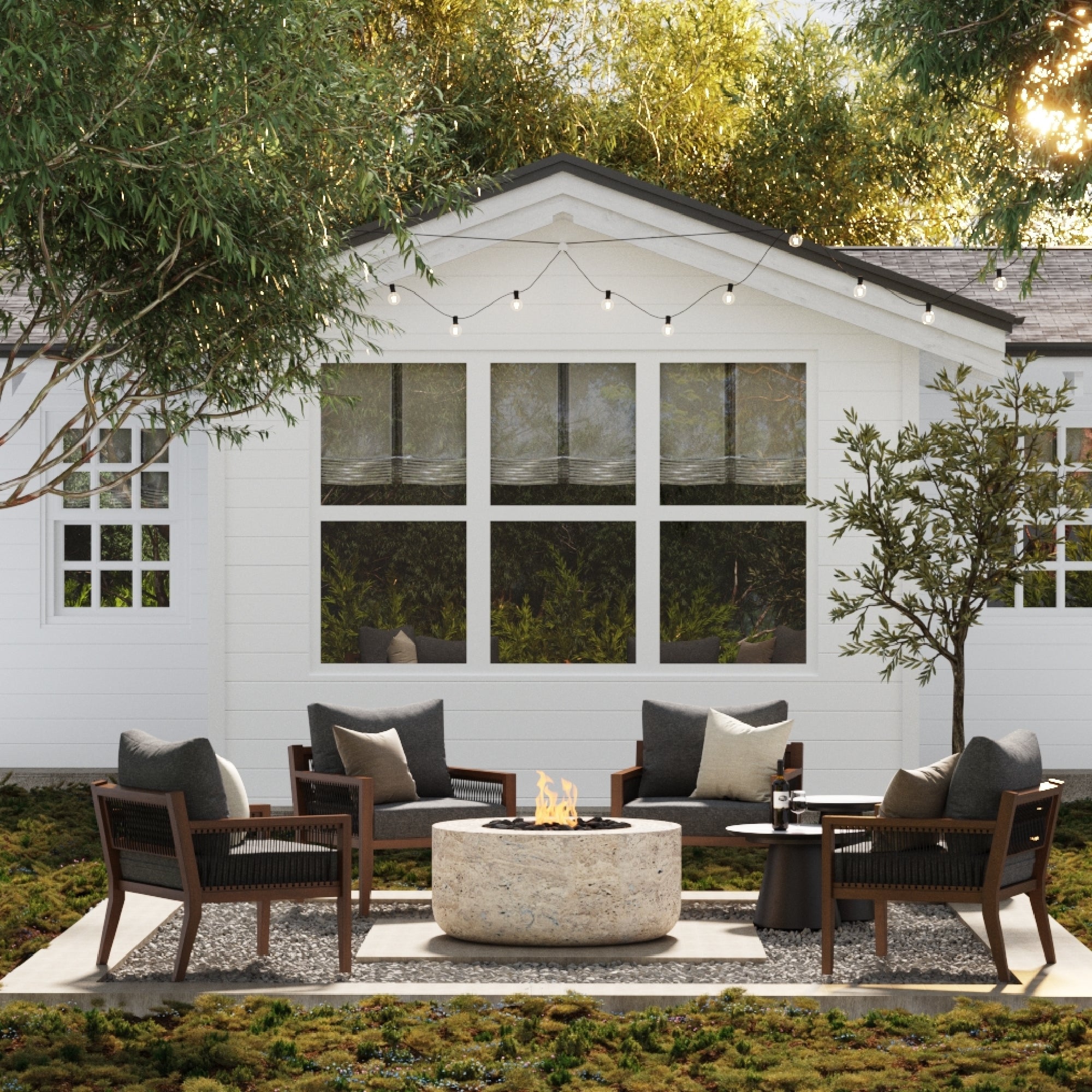 A cozy outdoor patio features four wooden chairs with dark cushions around a round stone fire pit, set on a patterned rug. String lights hang above, and the white house with large windows is surrounded by trees and greenery.