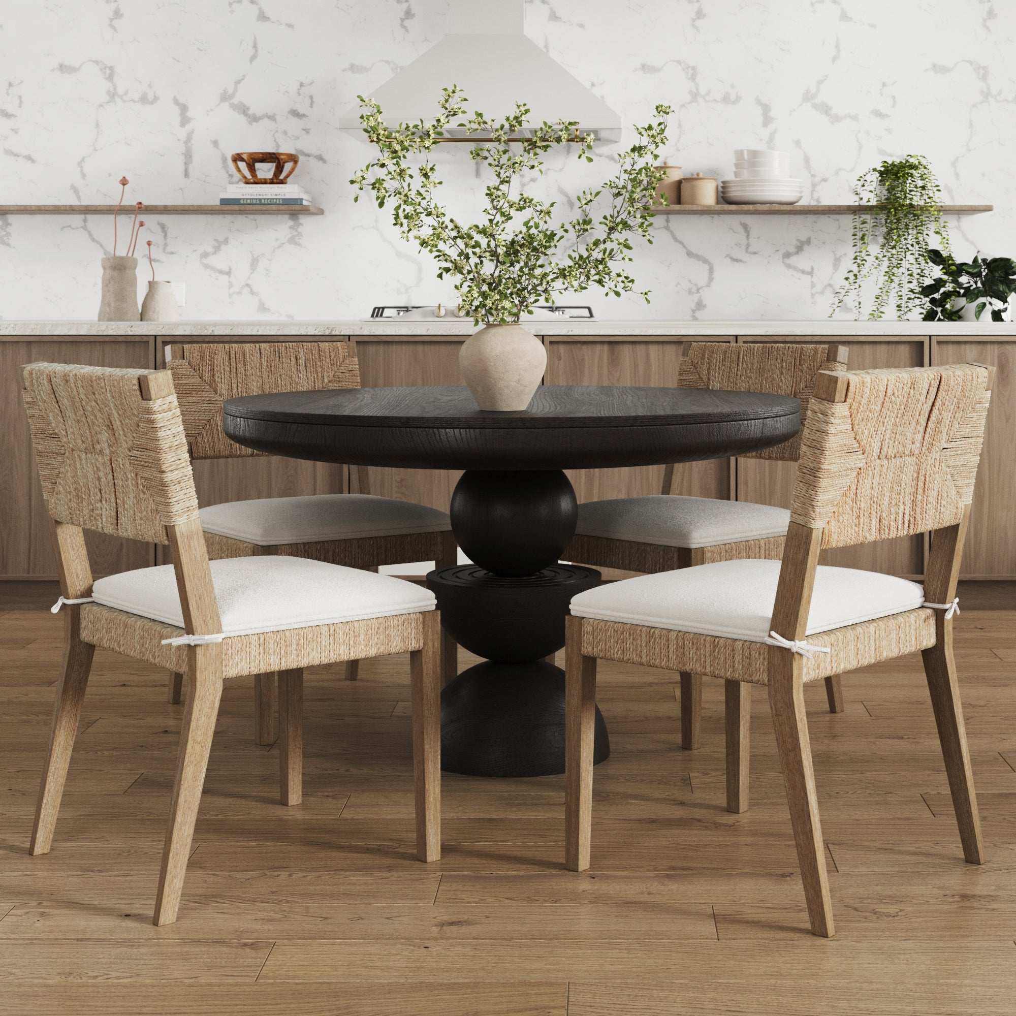 A modern kitchen features a round black dining table with a vase of green branches and four woven wooden chairs with white cushions, set on a light wood floor with minimalist shelves and neutral-toned decor in the background.