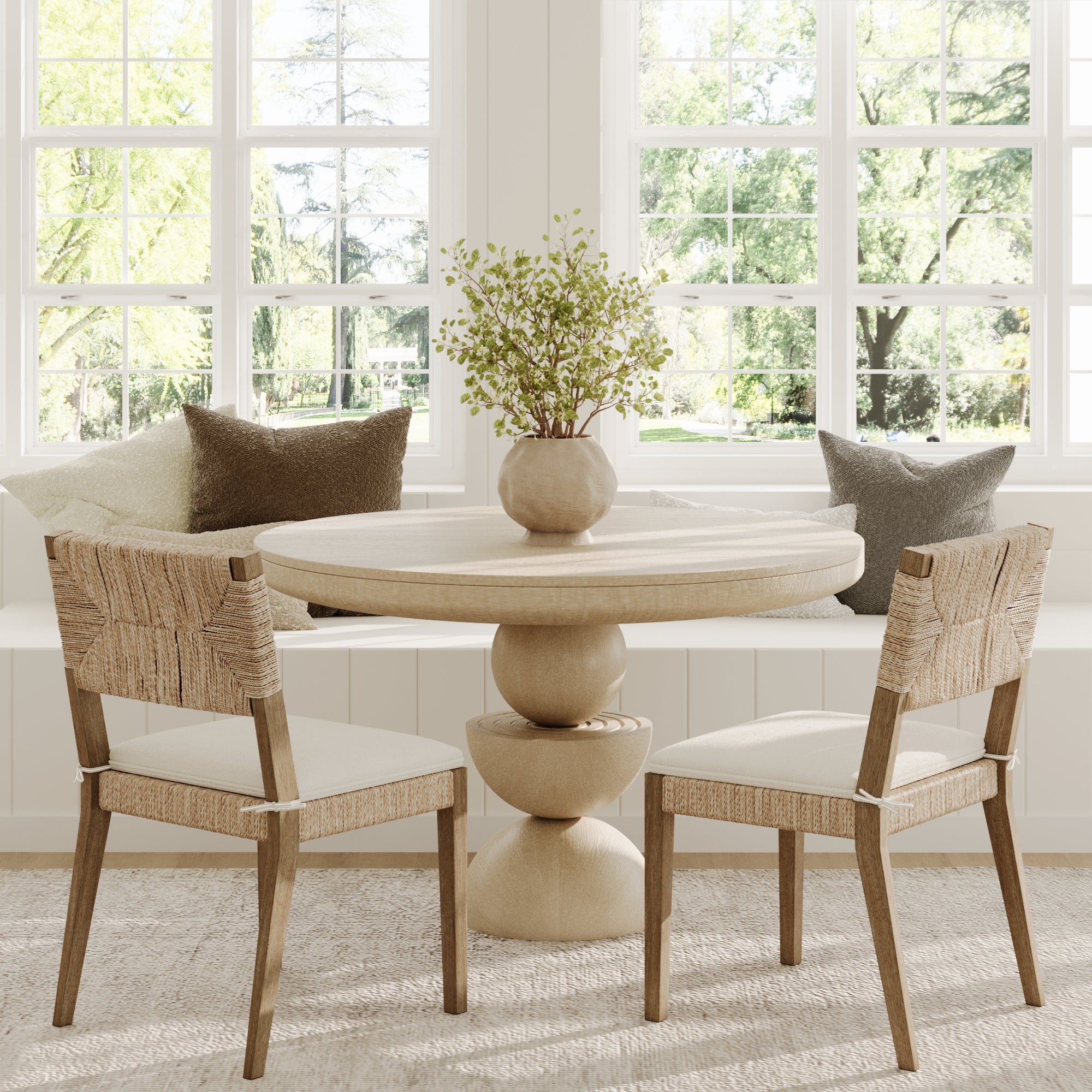 A cozy dining nook with a light wood round table, a solid wood bench with neutral pillows, and two Nathan James Boucle & Seagrass Dining Chairs in light brown set by large windows overlooking lush greenery.