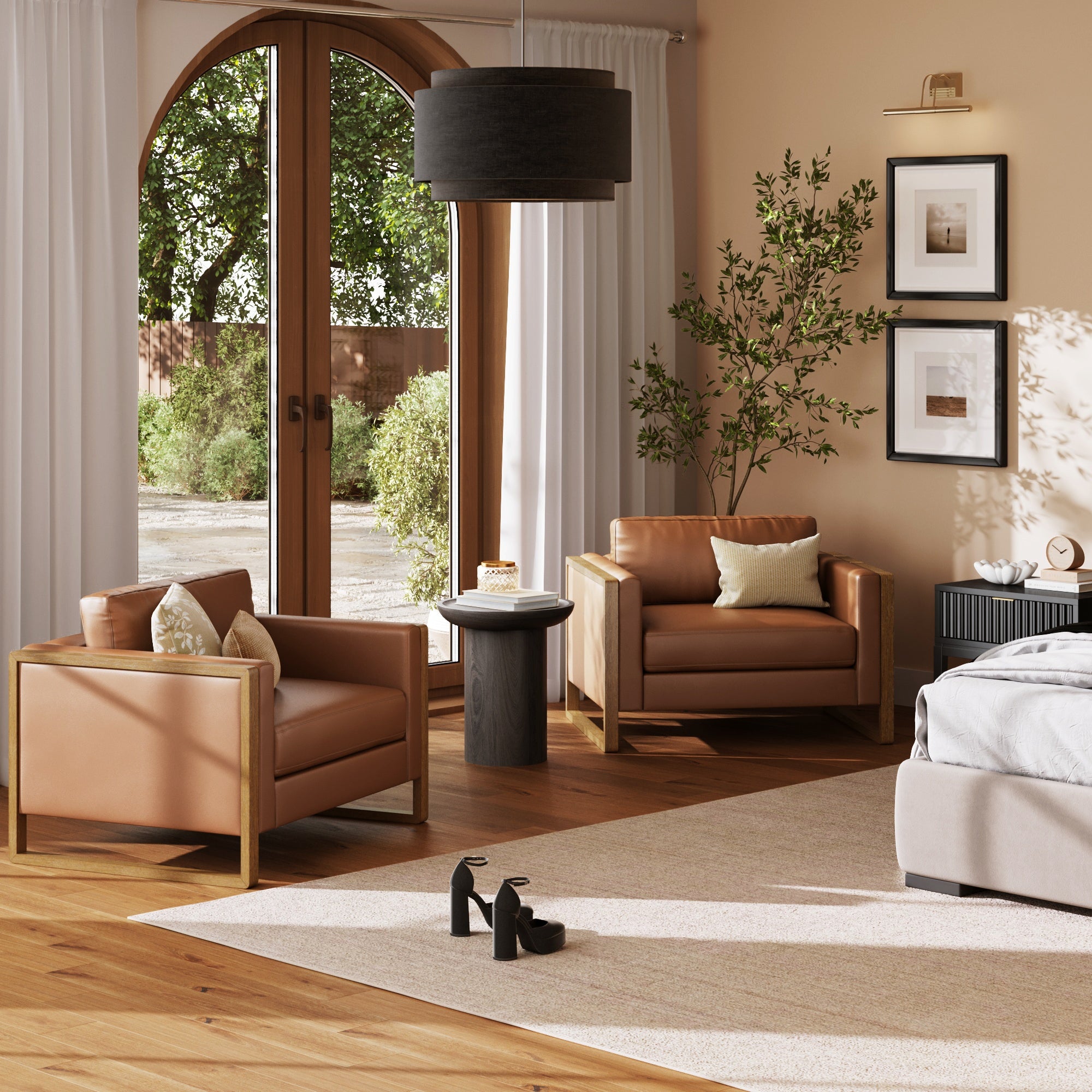 Two Nathan James Leather Modern Wood Frame Armchair Camel chairs sit in a cozy, sunlit room with wood flooring, a round black side table, a bed, framed art, an indoor plant, and black heels on a rug near large arched glass doors.