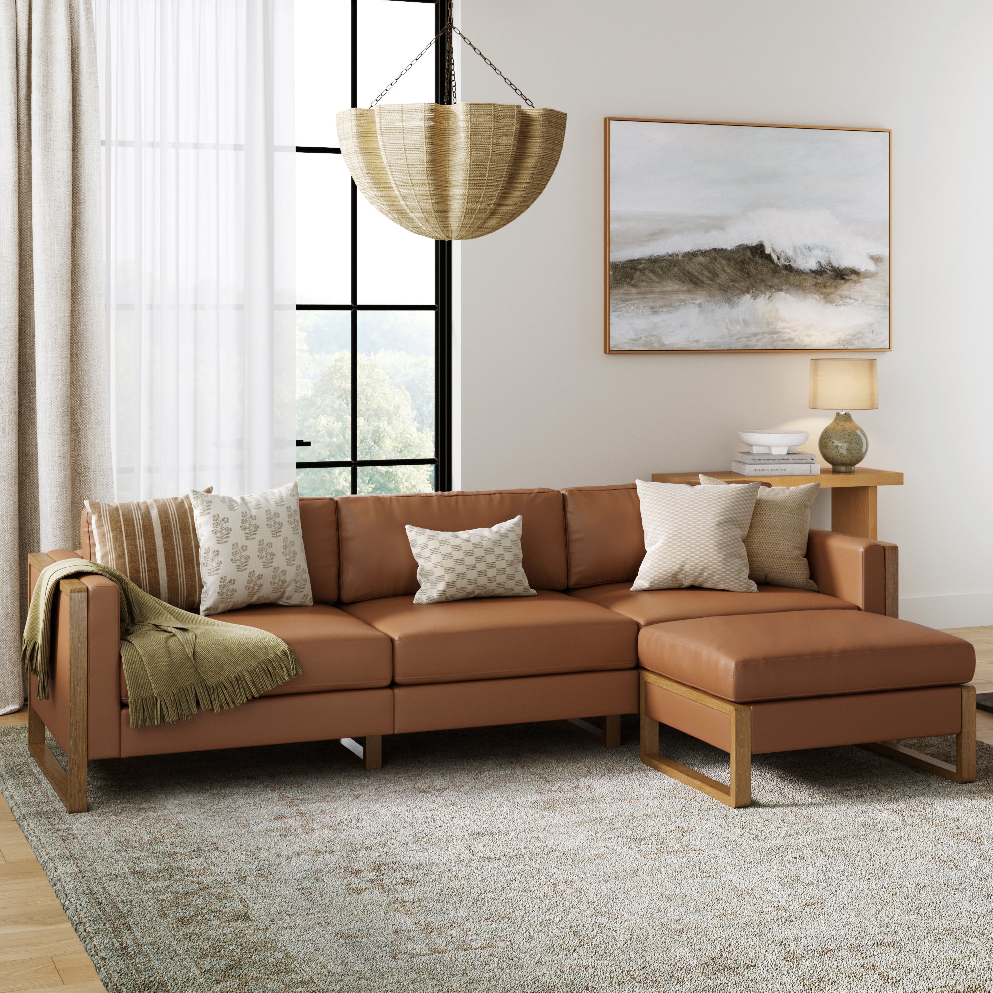 A modern living room featuring the Nathan James Upholstered Leather Sofa Standard Seat in camel, neutral throw pillows, a green blanket, wooden side table with lamp, large window, wave wall art, and a woven pendant light.