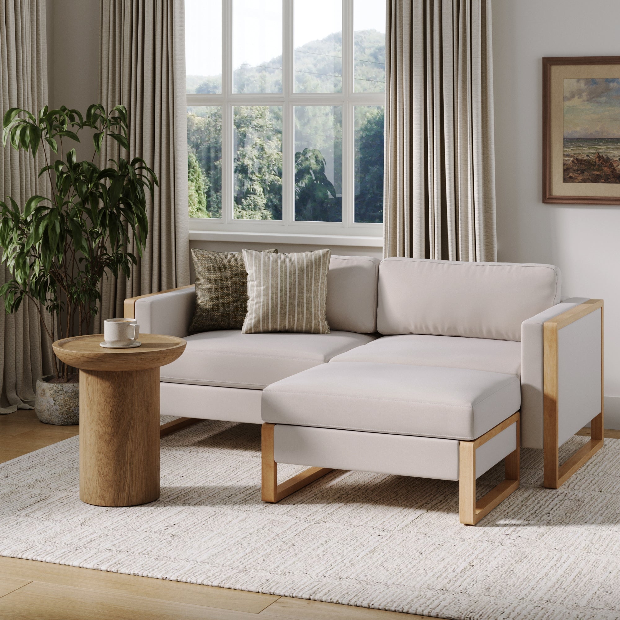 A modern living room with a Nathan James Soft Fabric Wood Frame Loveseat Sofa Couch & Storage Ottoman in bisque, paired with a round wooden side table holding a cup, a potted plant, and large curtained windows offering scenic outdoor views.